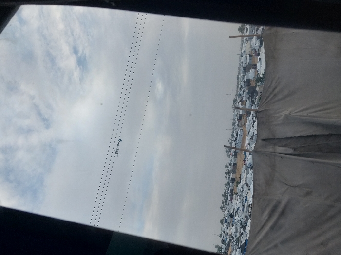 A view of a densely populated area, likely a refugee camp, from inside a structure. White tents or shelters stretch into the distance under an overcast sky. Power lines are visible across the sky. Part of a tent and a dark frame are in the foreground, suggesting the image was taken from within another shelter or vehicle. The landscape appears dry and crowded.