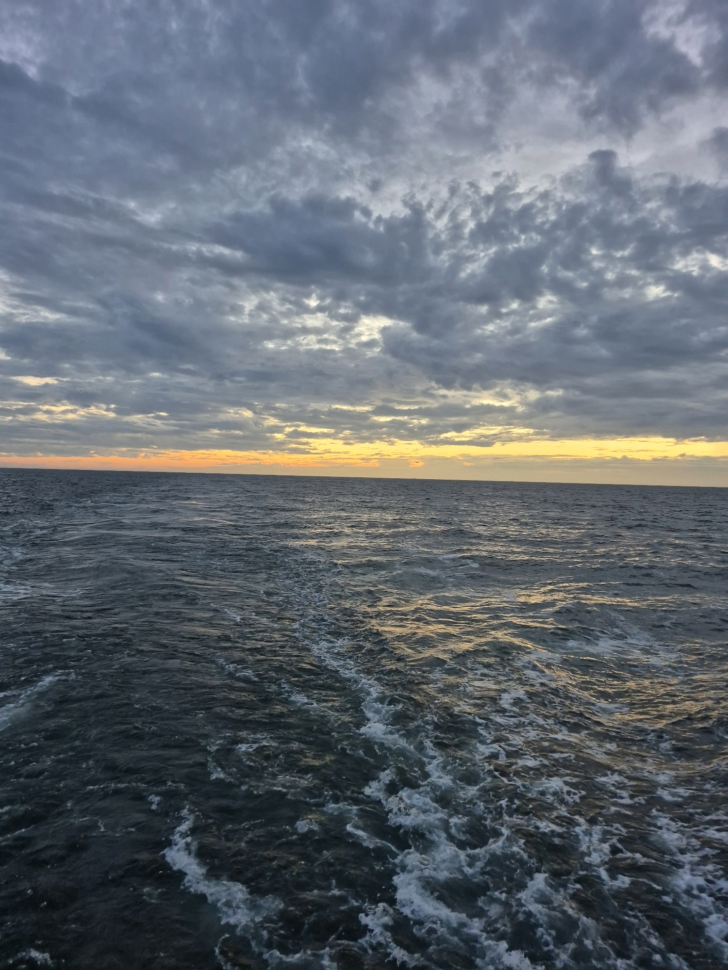 A wide-angle, eye-level shot captures a turbulent ocean scene under a dramatic, cloudy sky. The sky is filled with dark, gray clouds that are beginning to break apart, revealing a sliver of bright yellow and orange light near the horizon, suggesting a sunset or sunrise. The ocean itself is a deep, dark blue, with a noticeable wake trailing behind the viewpoint, indicating movement. White foam and churned water are visible in the wake, highlighting the choppy conditions. The horizon line is sharp and clearly separates the sea from the sky. The image does not contain any people or identifiable landmarks. The location is Unknown, Germany.