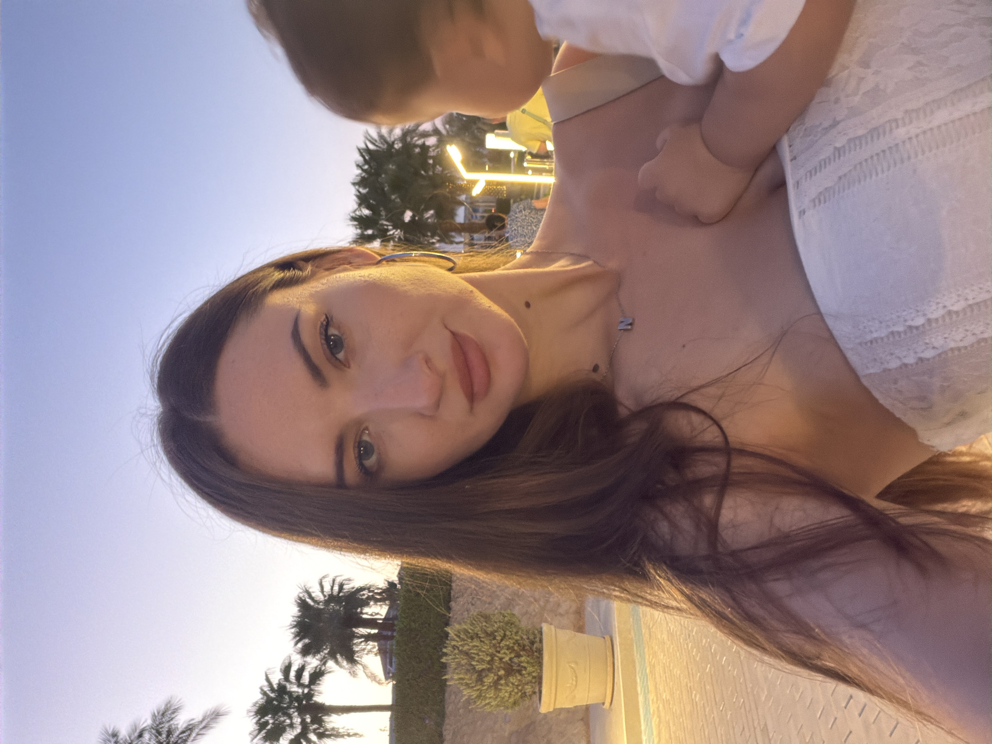 A medium shot selfie captures a woman holding a baby on a paved pathway at dusk in Sharm El Sheikh, Egypt. The woman, with long dark hair and blue eyes, is wearing a white lace top and a necklace with the letter "N". She is looking at the camera. The baby, held in her arms, has its back to the camera. Palm trees and illuminated lamps are visible in the background, along with other people in the distance. The lighting suggests a warm evening ambiance.