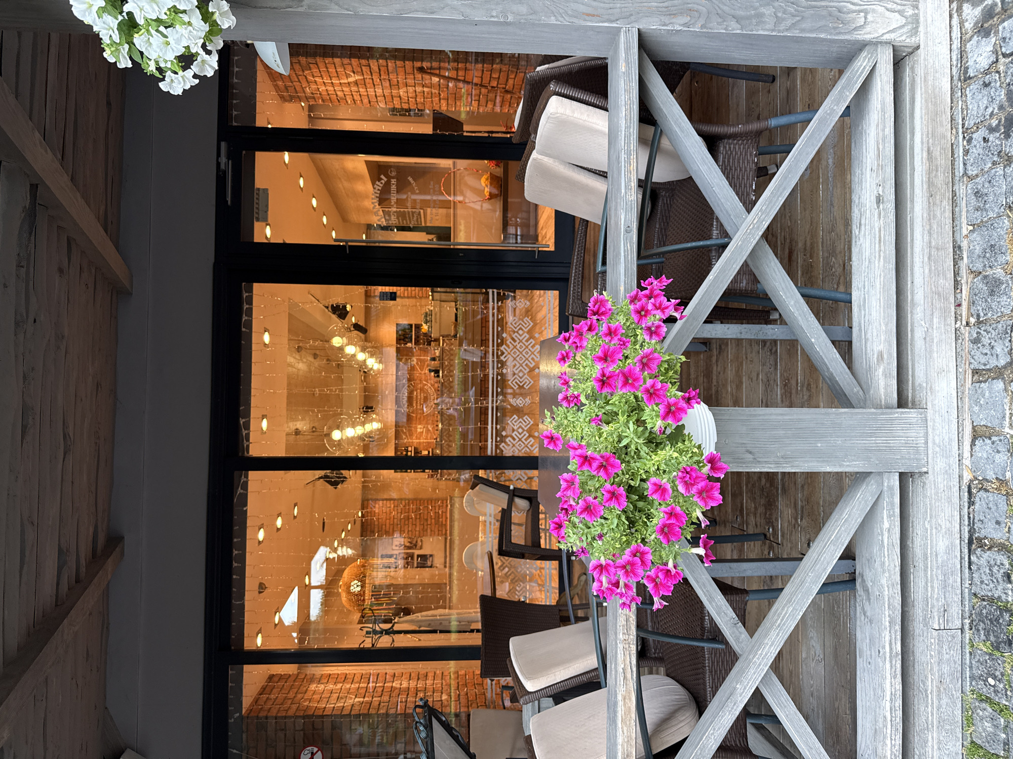 This image depicts an outdoor seating area of a cafe or restaurant located in the city of Yalta, Ukraine. The foreground is dominated by a wooden railing with a grey, weathered finish. A pot of vibrant pink petunias is placed on top of the railing, adding a splash of color to the scene. Behind the railing, there are several outdoor tables and chairs made of dark wicker material, some with light-colored cushions.
Through the large glass windows of the establishment, the interior is visible. The interior appears to be warmly lit with strings of fairy lights and round pendant lights. The walls are constructed of exposed red brick. Inside, there are more tables, chairs, and displays, suggesting a dining or retail space. One reflection in the window shows a basket filled with yellow fruits, possibly lemons.
The overall atmosphere is that of a European cafe, likely designed for outdoor dining or relaxation, situated in a city context, as indicated by the mention of Yalta.