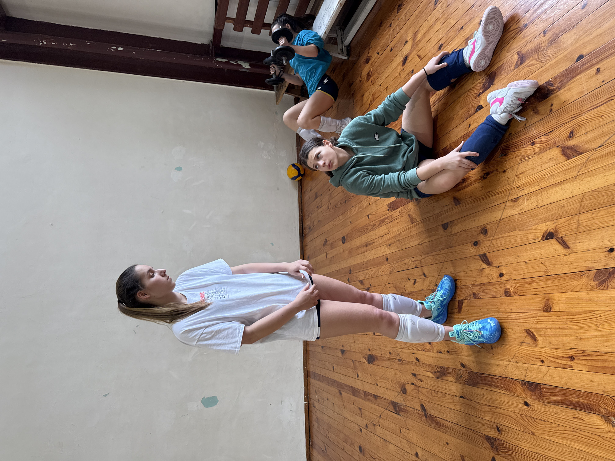 Three young women are in what appears to be a gymnasium or training room. The floor is made of wood planks. One woman is lying on her back, wearing a white t-shirt, white knee pads, black shorts, and blue sneakers. She is facing left. Another woman is sitting on the floor with her legs stretched out to either side. She is wearing a green sweatshirt, black shorts, dark blue leg warmers, and pink and white sneakers. She is looking at the camera. A third woman is in the background, holding two dumbbells and doing squats. She is wearing a blue t-shirt and black shorts. A volleyball is on the floor near the sitting woman. The location appears to be inside a building, but there is no definitive evidence to indicate it is in Sofia, Bulgaria.