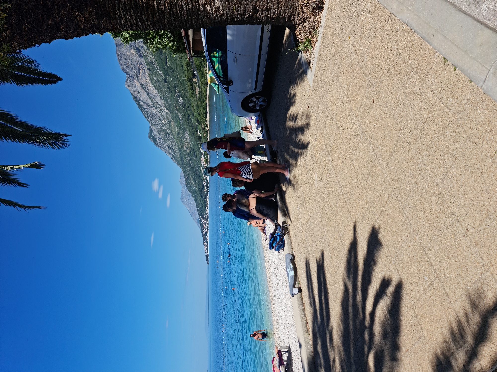 The image depicts a sunny day at a beach in Općina Gradac, Croatia. In the foreground, a paved walkway with palm tree shadows covers the ground. Several people are gathered on the beach and in the shallow water. A white car is parked on the right side of the walkway. The background features a coastline with a town nestled against a mountain range, all under a clear blue sky. The sea is calm and a barrier of buoys marks a swimming area.