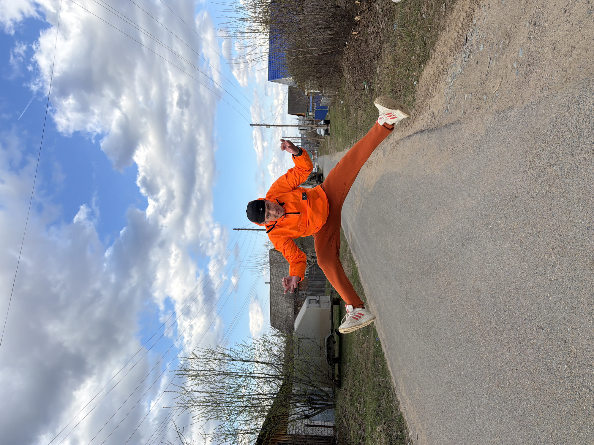 An outdoor, eye-level shot, originally rotated, depicts a young man in a dynamic, high-kick pose on a gravel road in Chaikovsky, Russia. He is positioned centrally, balancing on his right leg while his left leg is extended high in the air, creating a strong diagonal line across the frame. He wears a bright orange windbreaker, matching orange trousers, a black baseball cap, and white sneakers with red accents. His arms are bent, with his left hand open and his right hand pointing or gesturing, adding to the energetic feel. A subtle smile is visible on his face.
The setting is a rural or semi-rural street. The ground beneath him is a mix of asphalt and gravel, with dry grass and sparse, bare trees lining the edges of the road. In the background, simple wooden houses, some with blue roofs, are visible, typical of a village or small town. Utility poles with multiple power lines stretch across the sky, adding structural elements. The sky is partially cloudy, with large white and grey clouds against patches of bright blue, suggesting a clear but not entirely sunny day. A faint airplane contrail can be seen in the distance. The bare trees indicate either early spring or late autumn. The overall impression is one of youthful energy and playfulness captured in a moment of active movement.