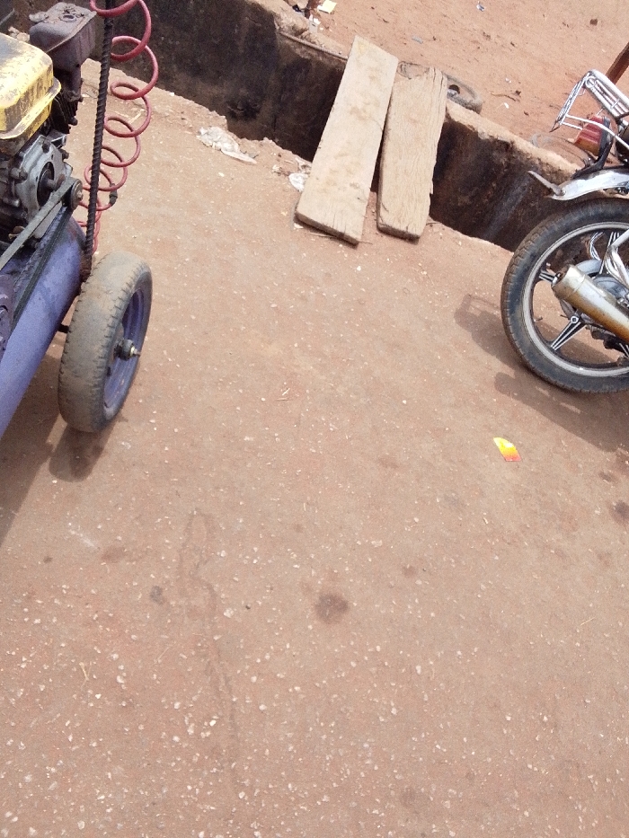 This image depicts a static scene in Ankpa, Nigeria, characterized by an unpaved or gravely reddish-brown ground surface.
In the left foreground, a portable air compressor is partially visible. It features a yellow-cased engine atop a blue or purple air tank, connected by a black V-belt. A single black rubber wheel with a purple hub is visible, supporting the unit. A red coiled air hose extends upwards from the compressor.
Towards the background, a dark, open drainage ditch or trench traverses the scene. Two light-colored, rectangular wooden planks are positioned vertically near the edge of this ditch, leaning against or resting upon its side. Behind these planks, additional debris and a dark tire are partially visible within the ditch area.
In the right foreground, the front wheel of a motorcycle is partially visible. The wheel has a black tire, a silver rim with black spokes, and a metallic exhaust pipe exhibiting some discoloration, positioned above the wheel. A chrome or silver metal rack structure is discernible behind the exhaust.
The reddish-brown ground is irregularly textured with scattered small white particulates and several dark, amorphous stains, possibly indicating oil or grease. A small, crumpled piece of orange and yellow plastic or paper litter is present on the ground near the motorcycle wheel. No individuals are depicted, and no specific actions or interactions are underway.