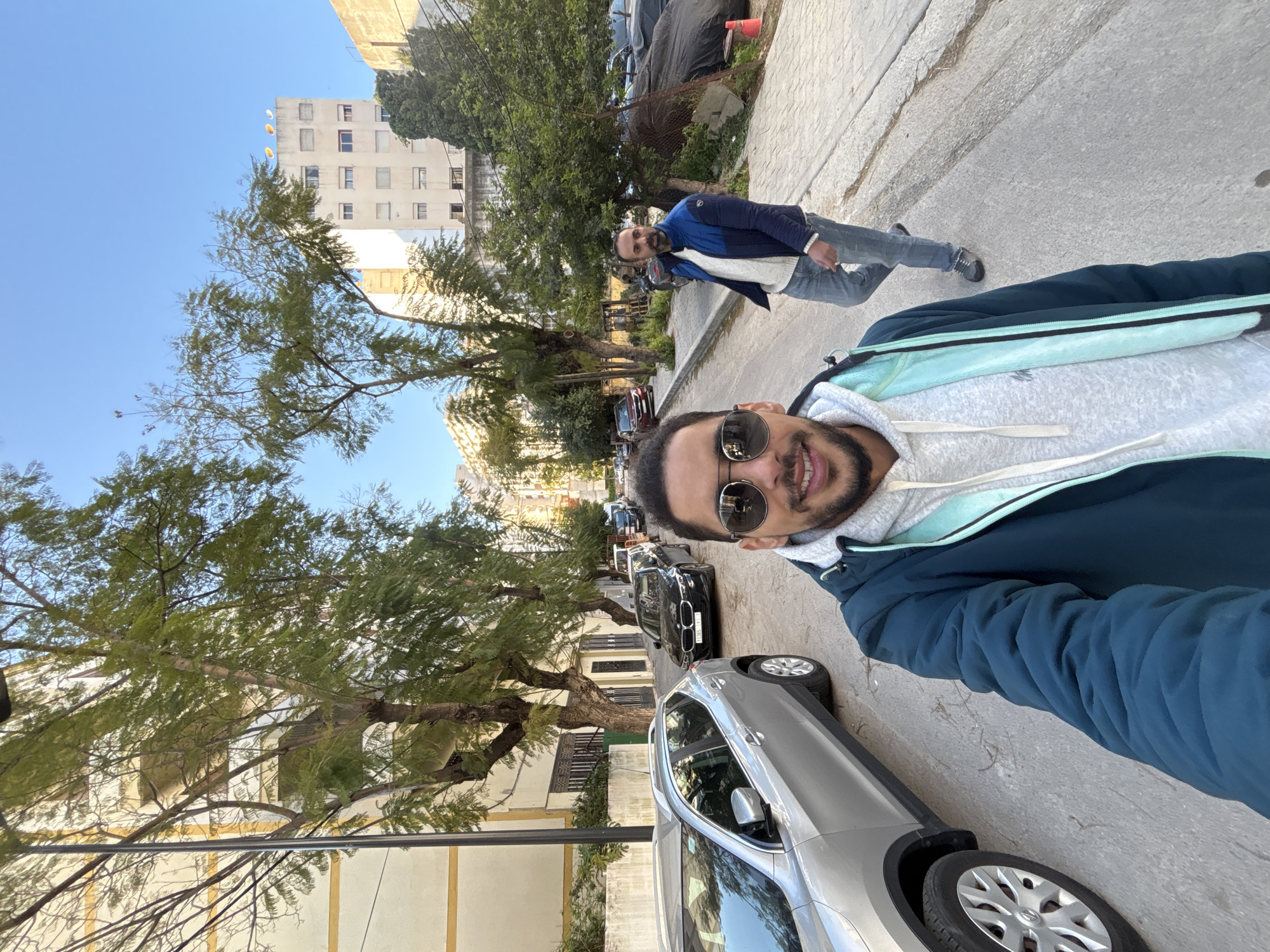 A selfie-style photograph captures two adult males on a paved city street in Tangier, Morocco, under a clear blue sky.
In the immediate foreground, facing the camera and positioned slightly left of center, is an adult male with dark hair, a beard, and dark sunglasses. He is smiling and wears a dark teal puffer jacket over a light grey hooded sweatshirt with light blue trim. His upper torso and head are visible.
Behind and to the right of the foreground subject, on the sidewalk, another adult male is walking towards the camera. He has dark hair and a beard, and is wearing a blue and black puffer jacket over a white sweater and blue jeans.
The street, which appears to be residential or mixed-use, runs into the background, lined with various parked vehicles. A silver SUV is partially visible on the left, next to the foreground subject. Further down the street, a black BMW sedan and other cars are parked on the right side.
Buildings of varying heights and architectural styles line both sides of the street. On the left, a multi-story building with cream-colored walls and yellow horizontal trim is visible. On the right, a taller, plainer white building with numerous windows and satellite dishes on its rooftop is visible in the distance. The street is also lined with numerous mature trees featuring green foliage. On the right sidewalk, near the walking individual, there is a black tarp covering an object, with a small orange cone nearby. A dark street lamp post is visible on the far left. The lighting suggests daytime conditions with a clear sky.