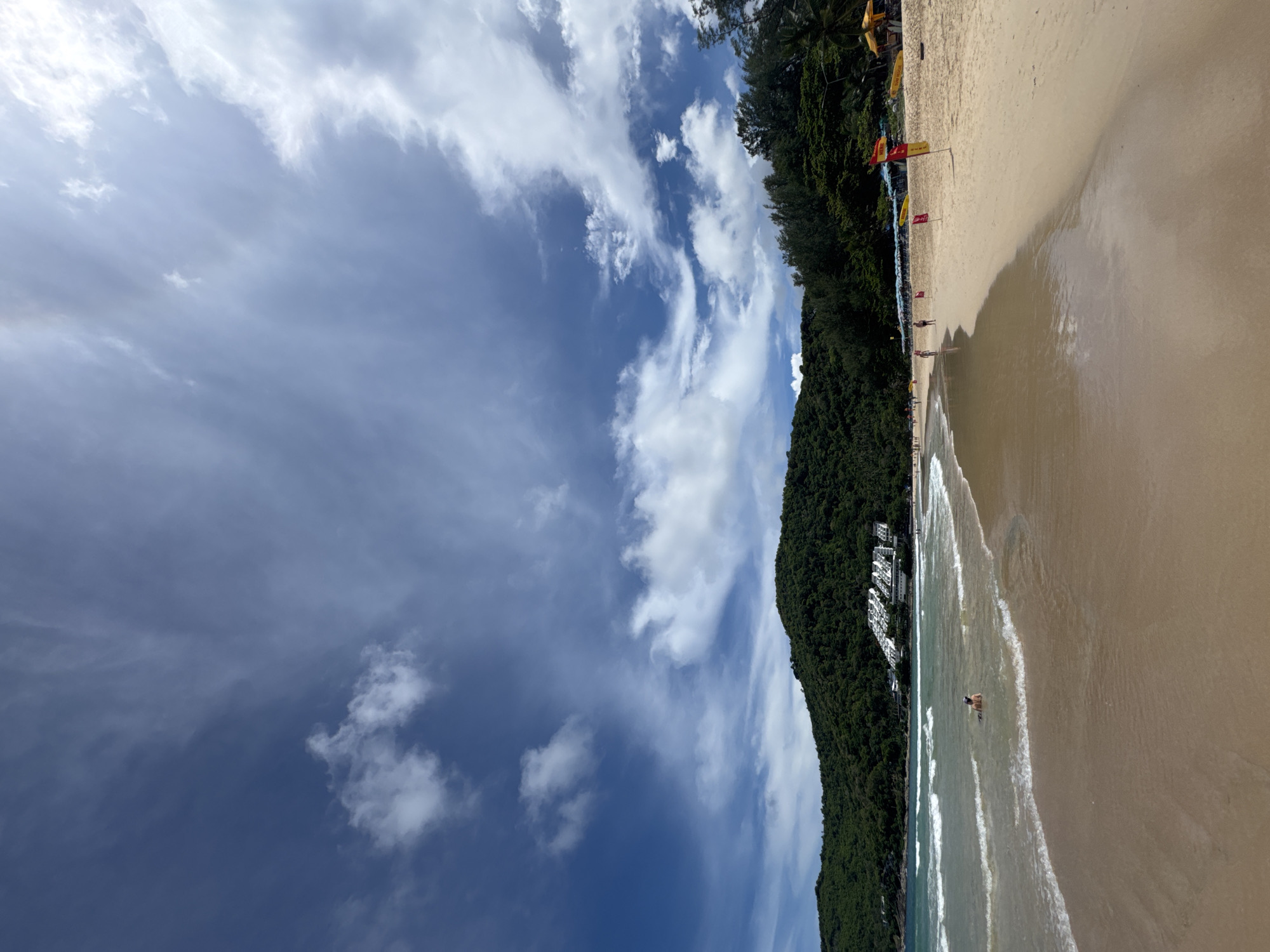 The image depicts a wide, sandy beach in Rawai, Thailand, under a partly cloudy sky with dramatic cloud formations. Gentle waves lap the shore, creating a foamy edge on the wet sand. In the shallow water, a single person is partially submerged, appearing to relax or swim. The beach is lined with lounge chairs and umbrellas, suggesting a resort area. Red and yellow flags, possibly indicating a designated swimming area or surf rescue station, are visible on the sand. Behind the beach, a lush, green, tree-covered hill rises, with a cluster of white buildings visible partway up its slope. The overall scene conveys a peaceful and scenic beach environment on a bright day.