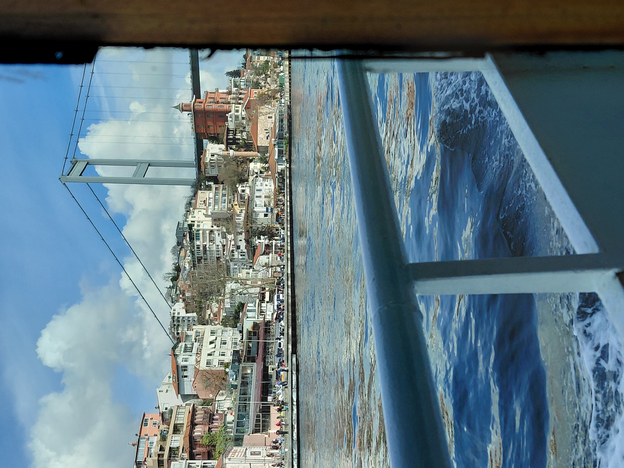 The image shows a view from a boat sailing on the Bosphorus in Sarıyer, Turkey. The cityscape of Sarıyer is visible, featuring multi-story buildings of varying architectural styles, including one prominent red brick building with a tower. A suspension bridge spans the waterway, its support structure visible against a blue sky with scattered white clouds. The foreground includes the boat's railing and the water, agitated and frothy, suggesting the boat is in motion.