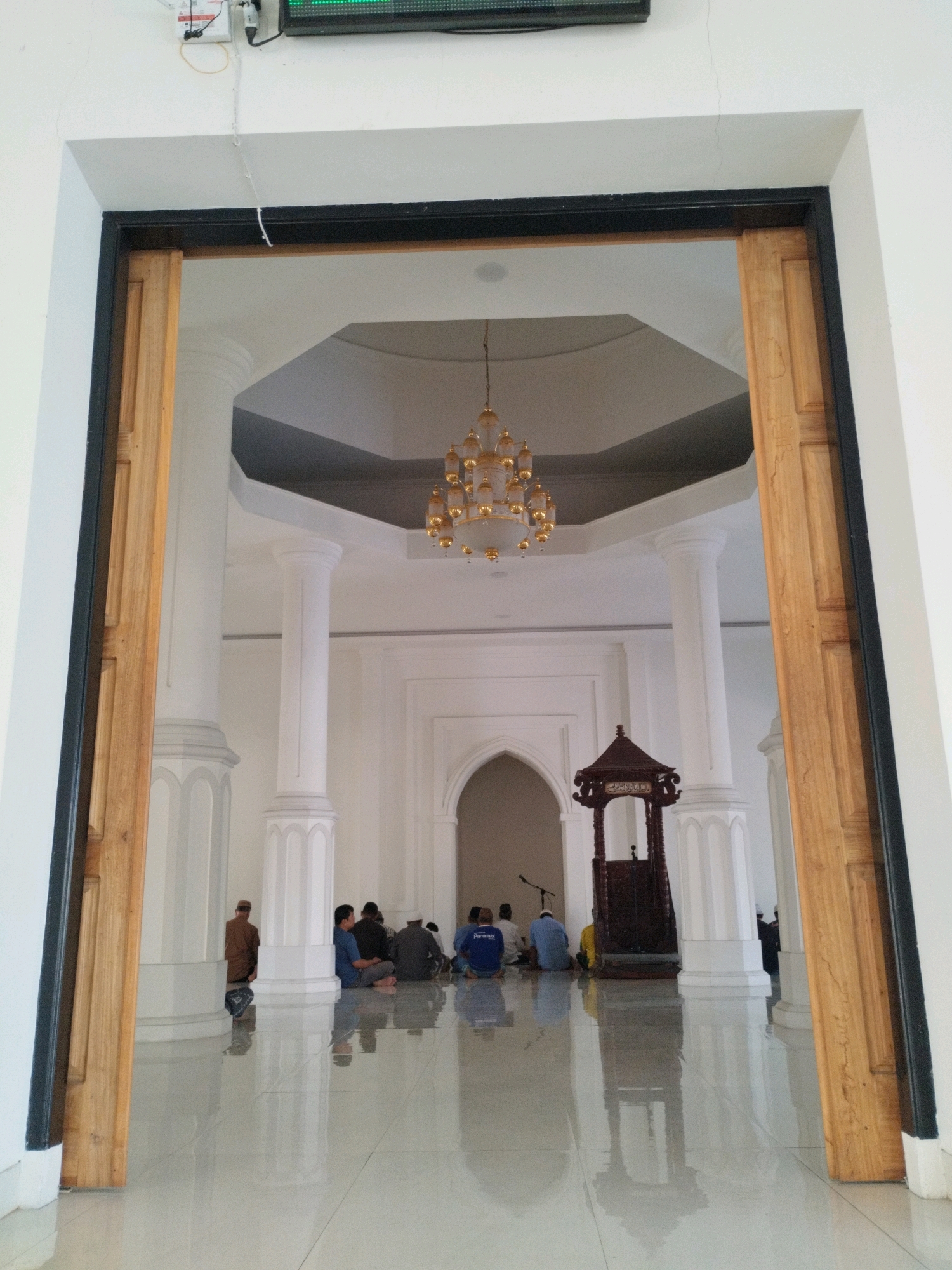 This image shows the interior of a mosque, viewed through an open doorway with wooden doors on either side. The mosque features a high, octagonal ceiling with a decorative chandelier. Large, white pillars support the ceiling. In the center of the visible area, a dark wooden pulpit stands. Several people are seated on the white, reflective floor, facing towards the pulpit, some in prayer. The architectural style and the presence of a pulpit suggest this is a place of Islamic worship.
While the image itself does not explicitly state its location, the request specifies that it is in the city of Gorontalo, Indonesia. Therefore, this scene is depicted within a mosque in Gorontalo, Indonesia. The environment is clean and well-lit, with a focus on the architectural elements and the spiritual activity taking place.