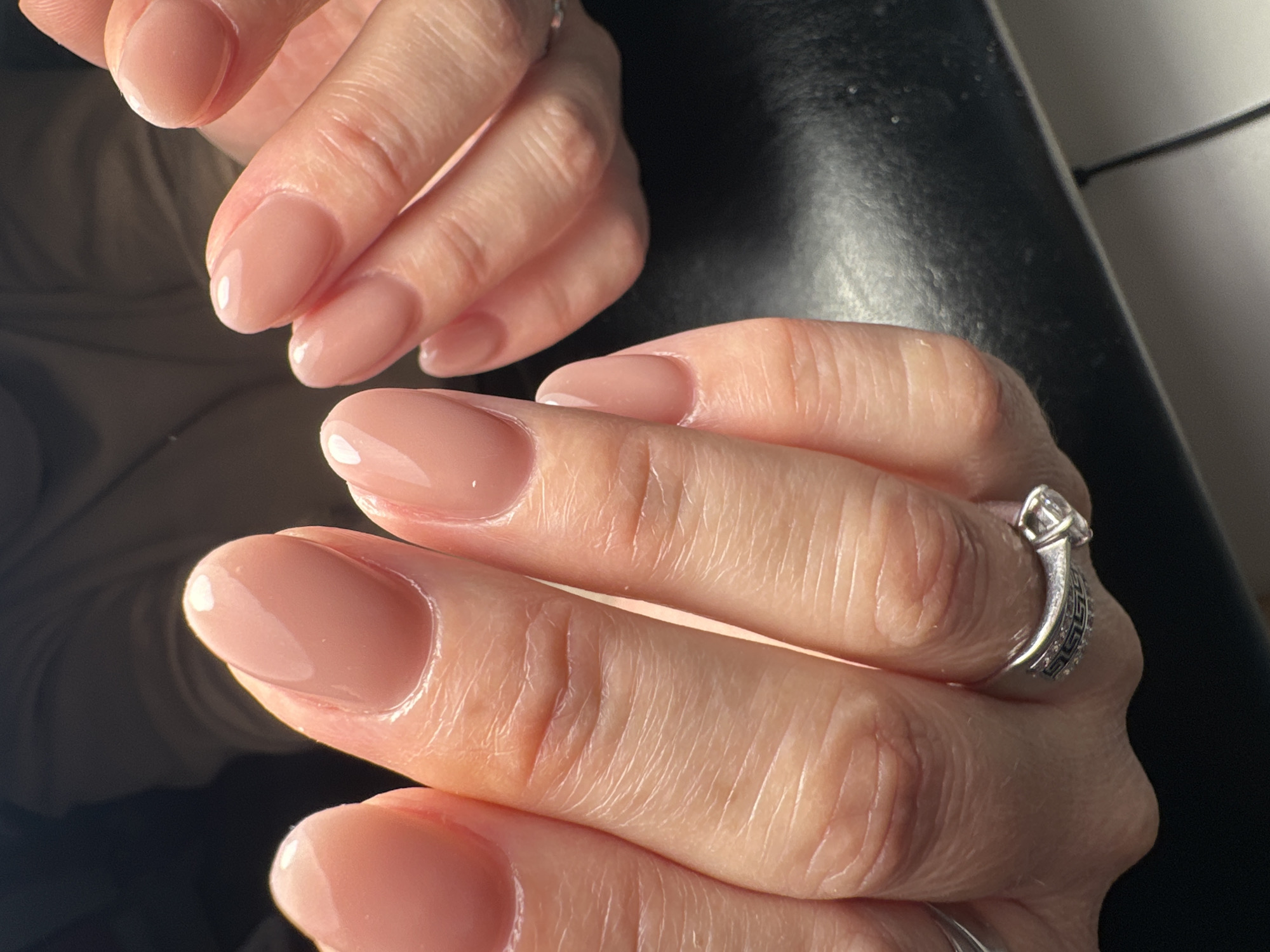 The image shows two hands with manicured fingernails painted in a nude shade. The nails are shaped in an oval or almond style. A ring is visible on one of the fingers. The background is blurry but appears to be a mix of dark and light tones. The image does not include sufficient information to determine the location in Chaikovsky, Russia. There is no information on people, subjects, objects, actions, interactions, and the environment other than the description above.