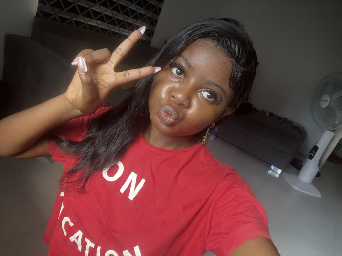 The image displays a close-up, selfie-style photograph of a single female subject, positioned indoors in Badore, Nigeria. The subject, a young adult with dark skin and long dark hair styled to the side, is wearing a red t-shirt with white text partially visible, displaying "ON" and "CATION." She has a small silver stud piercing in her left nostril and a gold-colored cross earring in her right ear. Her lips are pursed in a "duck face" expression. Her left hand is raised, forming a "peace sign" gesture with two fingers extended; her fingernails are long, almond-shaped, and appear to be painted a light, possibly iridescent color. The background consists of a dark furniture item and a patterned wall section to the subject's right, and a white standing electric fan to her left. The floor is a light tone. The scene is illuminated by interior lighting.