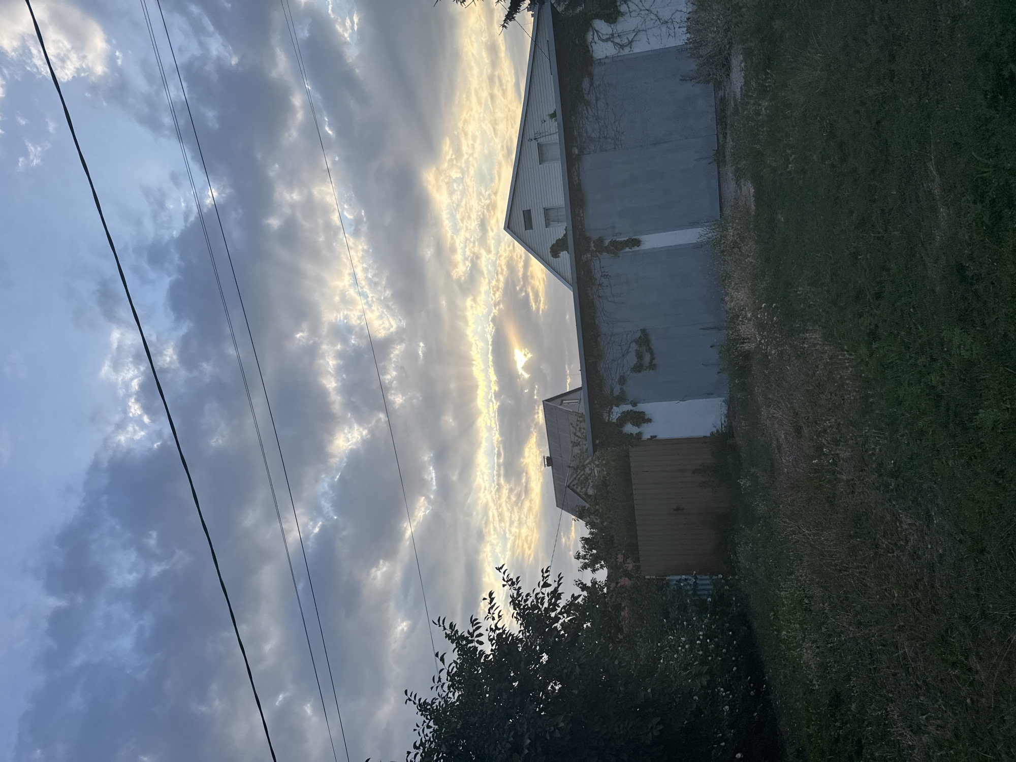 An outdoor, low-angle, vertical shot captures a scene in Zaporizhzhia, Ukraine, likely during dawn or dusk. The sky is filled with a dramatic display of grey and white clouds, with streaks of sunlight piercing through, creating crepuscular rays. Power lines are visible crisscrossing the sky. In the middle ground, a grey garage with a corrugated metal door is prominent, with vines growing on its surface. Behind the garage, a white house with a gabled roof and two windows is visible. To the left, a tall tree with dark green foliage stands in front of a wooden fence. The foreground is a dark, grassy area, appearing overgrown and unkempt. The overall impression is of a slightly weathered, rural or suburban setting under a dynamic sky.