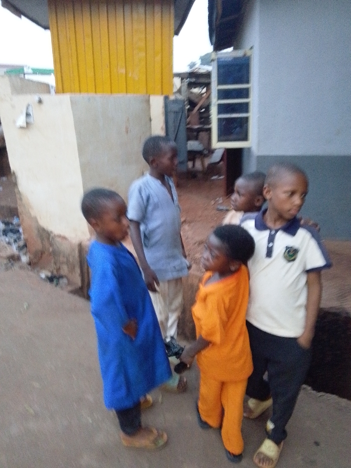 Five young children are standing close together outdoors. One wears a long blue robe with dark leggings and tan sandals. Another is dressed in a light gray long-sleeved shirt and beige pants. A child is in full orange, including shoes. The last wears a white shirt with dark blue accents and black pants. Buildings of varied materials are in the background, including one with a yellow section at the top.