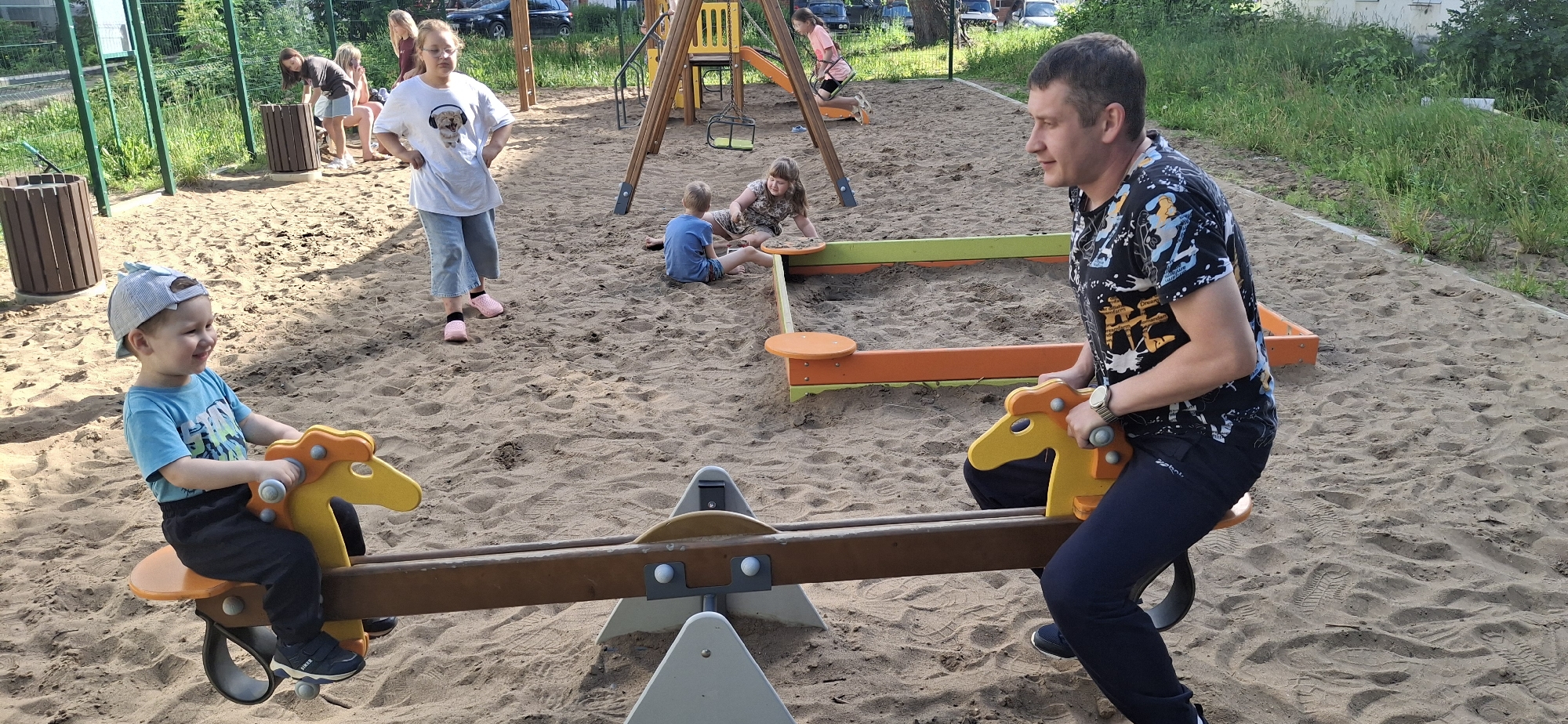 In the city of Chaikovsky, Russia, a public playground is depicted with sandy ground and sparse vegetation. In the foreground, a man in a black t-shirt and dark pants is seated on a seesaw, his left arm resting on the yellow horse-shaped handle. To his left, a young boy in a blue t-shirt and a grey cap is also on the seesaw, facing left and smiling. He is holding onto the yellow horse-shaped handle with both hands. In the mid-ground, a sandbox with orange and green trim is visible. Two girls are near the sandbox; one in a blue t-shirt is sitting with her legs extended, and another in a patterned dress is kneeling. Further back, a young girl is on a swing set. To the left of the frame, two women are standing, one in a white t-shirt with a dog graphic and blue jeans, and another in a dark t-shirt and skirt. Behind them, a wooden trash bin is situated near a green fence. The overall scene captures a moment of leisure and play on a sunny day.