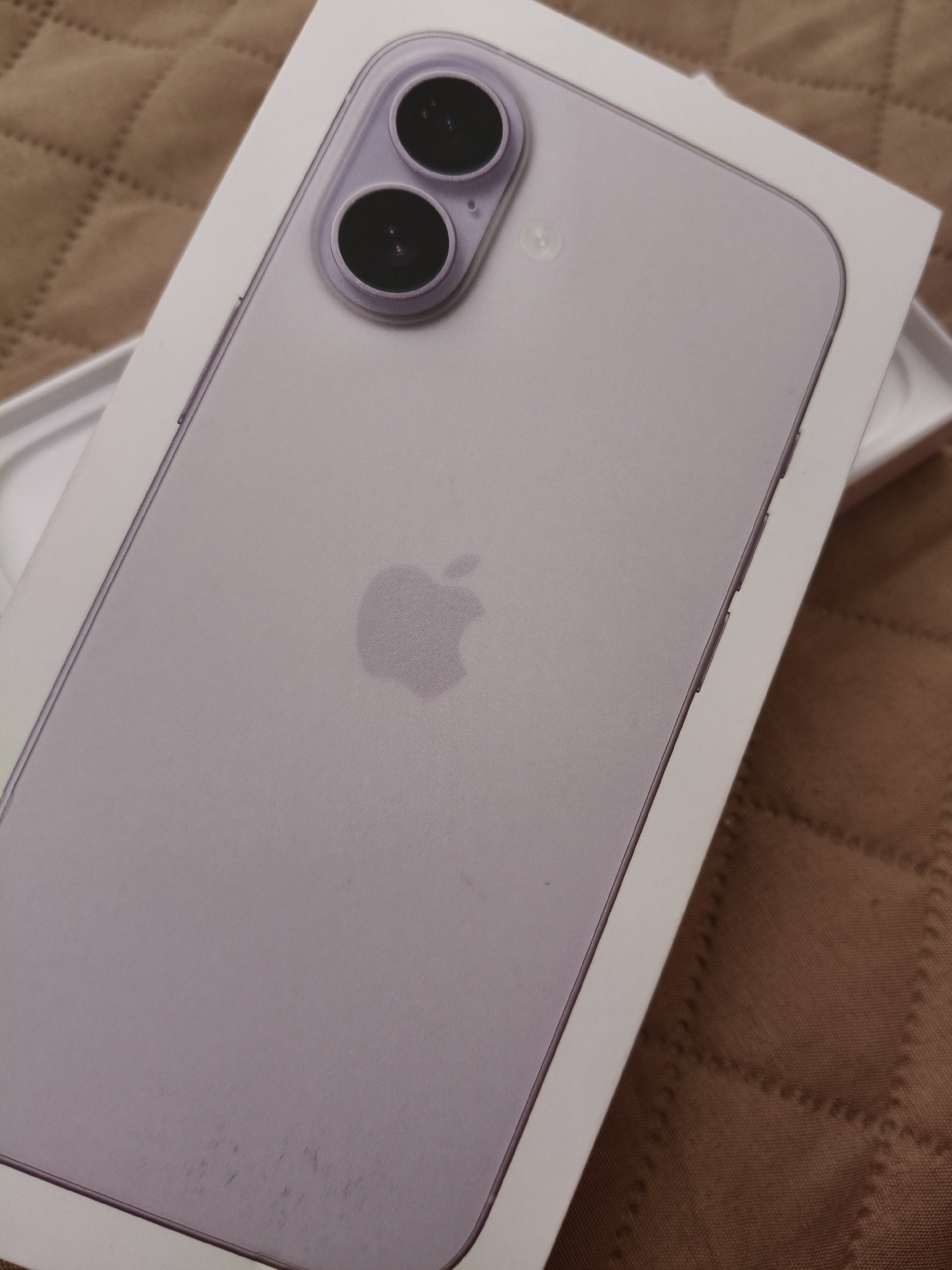 The image shows the back of a lilac-colored iPhone in its original white packaging. The phone has a dual camera system in the upper left corner and the Apple logo in the center. The background is a brown quilted material. The location in Rakitovo, Bulgaria is not evident from the image.