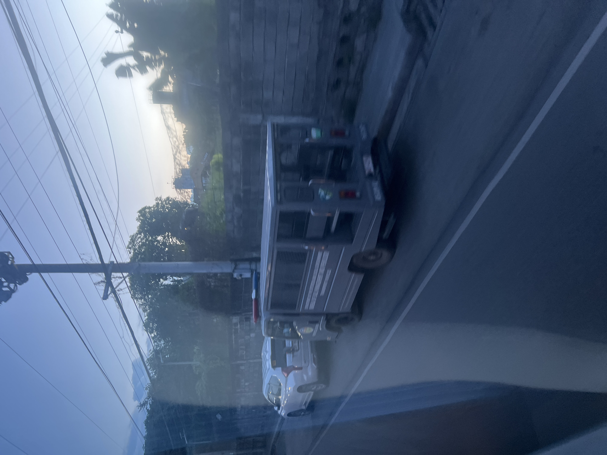 The image shows a street scene at dusk in Davao City, Philippines. Two vehicles are visible on the road: a white SUV on the left and a gray utility vehicle, possibly a police van or a similar government vehicle, on the right. The utility vehicle has flashing red and blue lights on its roof and appears to be stationary or moving slowly. The SUV is behind it, also appearing to be moving slowly or stopped. The road is paved, and there is a concrete wall with some vegetation growing on top along the right side of the street. Overhead power lines crisscross the sky, which is a pale blue with some clouds and hints of sunset colors. In the background, trees and some buildings are visible, suggesting an urban or suburban environment. The overall lighting indicates it is either sunrise or sunset.