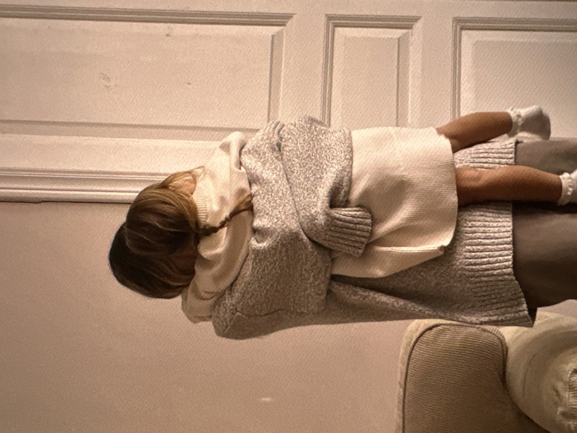 A young person rests horizontally on a couch or soft seating arrangement, facing away from the camera. They are wearing white socks and a light-colored sweater. They are covered by a grey knitted cardigan. The background features a wall with horizontal, rectangular paneling. The location is undetermined, and there's no information to suggest it is Yerevan, Armenia.