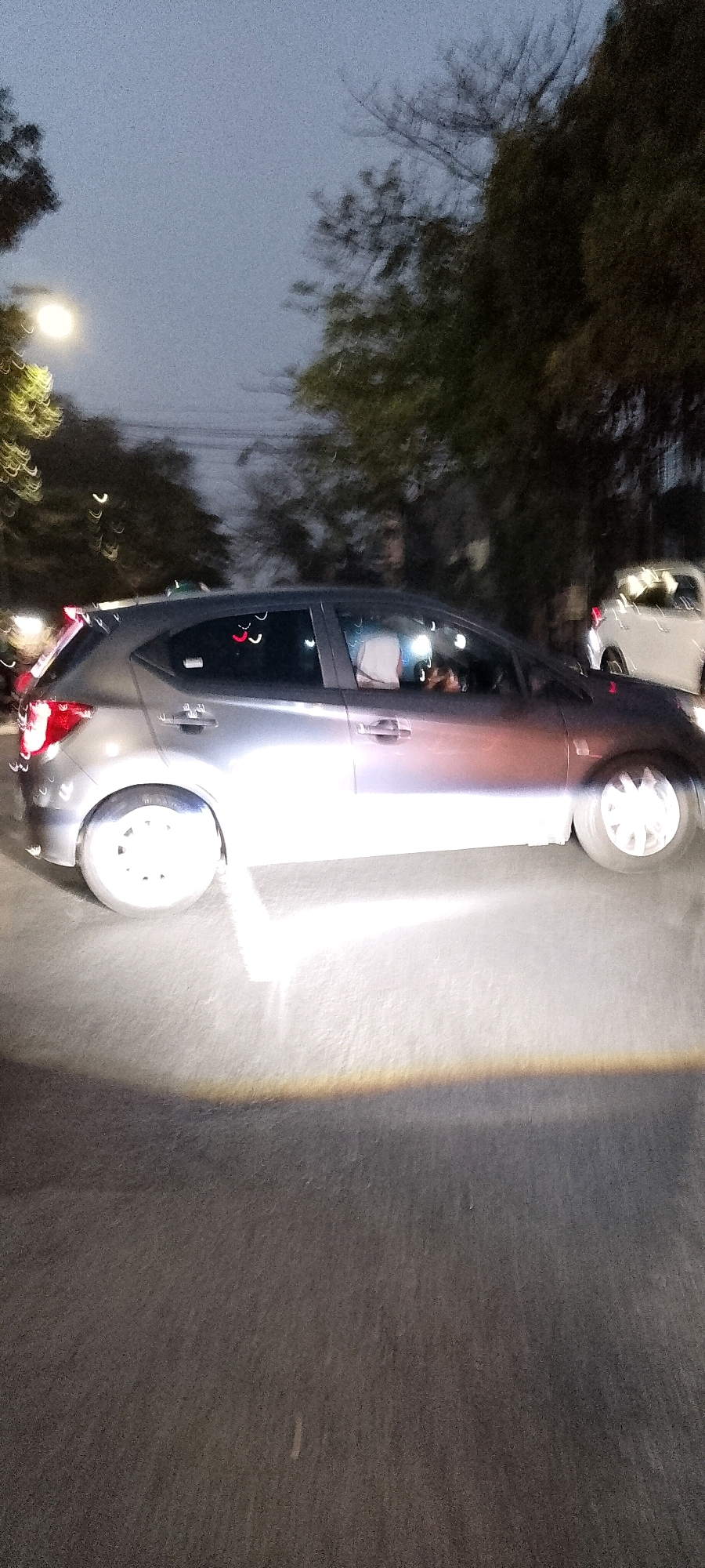 A silver hatchback car is parked on a paved road at night, with trees and streetlights in the background. The car's headlights are on, illuminating the road in front of it with a bright white light. The car's windows are tinted, and it is difficult to see inside. There is another white car in the background, also with its headlights on. The sky is dark and overcast, and there are some blurred lights in the distance, possibly from other vehicles or buildings. The scene appears to be in a suburban area. Based on the presence of streetlights, trees, and paved roads, this could be a location within the city of Depok, Indonesia.