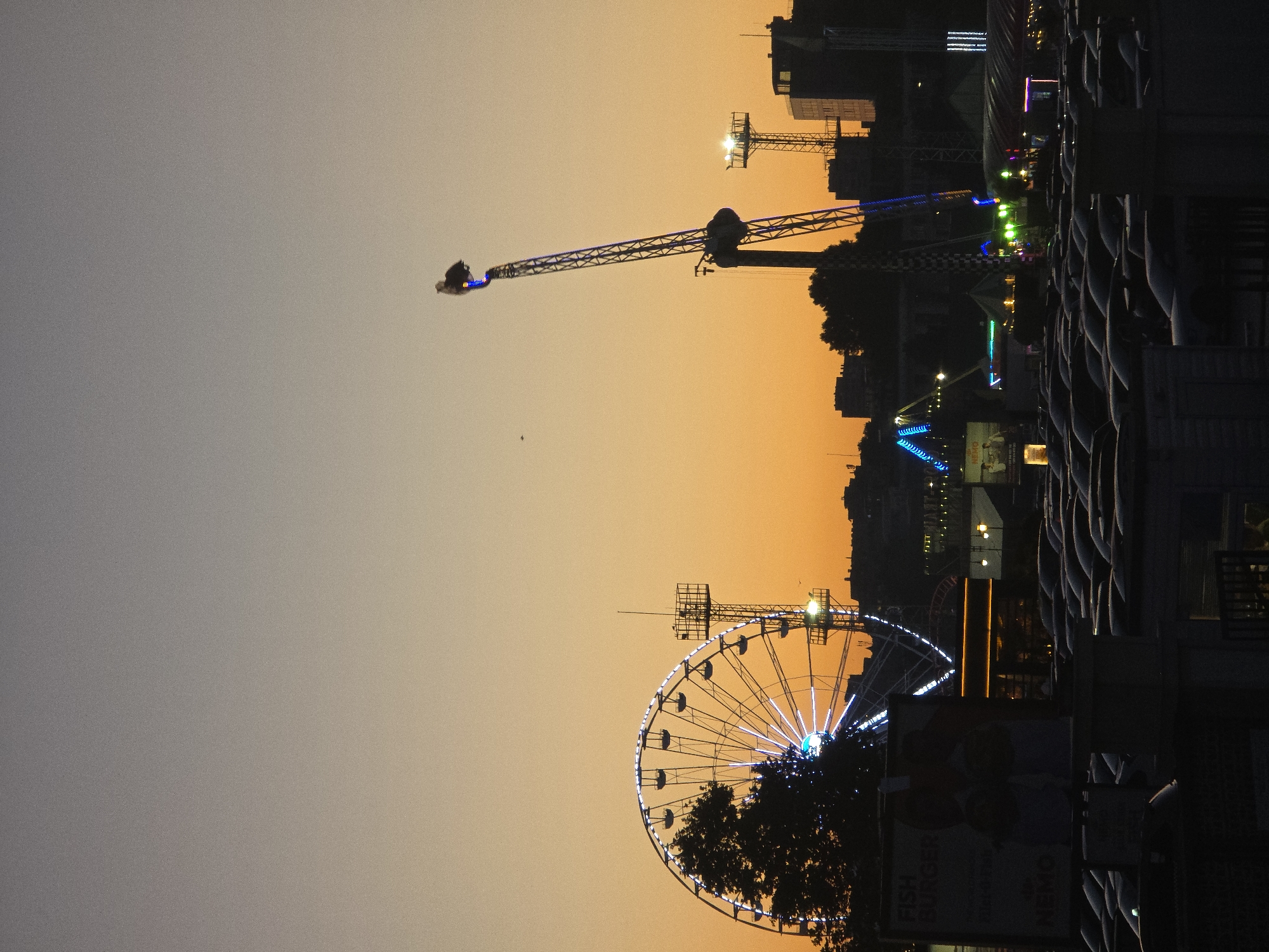 The image depicts an outdoor amusement park scene at dusk in Varna, Bulgaria. In the foreground, a dark silhouette of a parking lot filled with cars suggests a public gathering. A prominent Ferris wheel, illuminated with white lights, is visible on the left, partially obscured by the dark silhouette of a tree. To the right of the Ferris wheel, a tall amusement ride with a tilting mechanism is silhouetted against the warm orange and grey gradient of the sunset sky. Various other amusement rides, illuminated by colorful lights, are scattered throughout the park in the background. Buildings and structures, including what appear to be more amusement park attractions and possibly permanent city buildings, are visible on the horizon. The overall atmosphere is that of a lively evening at an amusement park, with the fading light of day contrasting with the artificial illumination of the rides.