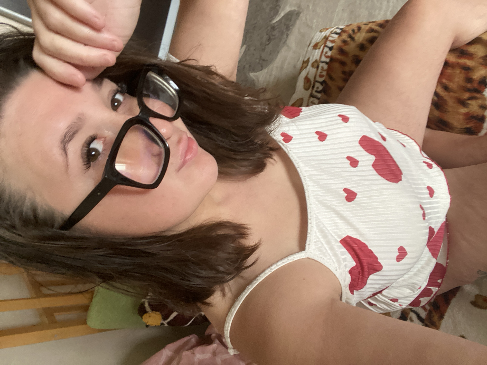 The image shows a young woman wearing glasses and a white camisole with red hearts. She is indoors, lying on a bed. The background includes a wooden headboard and a pillow with a leopard print. The image does not provide any information about the location in Chaikovsky, Russia.