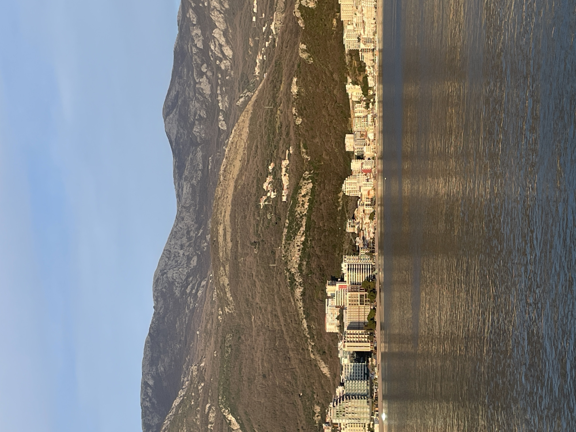 The image displays a panoramic view of the coastline of Budva, Montenegro, observed from the sea. The foreground consists of a calm body of water, likely the Adriatic Sea, exhibiting subtle ripples and reflections of the distant cityscape and sky.
The midground features the urbanized coastline of Budva. A continuous line of multi-story buildings, including modern hotels and apartment complexes with varied architectural styles (some featuring light-colored facades, others with glass elements), lines the waterfront. A narrow, light-colored beach is visible directly in front of these structures, with sparse vegetation, possibly trees or shrubs, interspersed along the shoreline. No discernible individuals or specific actions are identifiable at this distance.
The background is dominated by a range of large, rugged mountains rising immediately behind the city. The mountain slopes are covered with sparse, dark vegetation, including patches of deciduous trees or scrub, and prominent rocky outcrops. Scattered smaller residential buildings or villas are visible nestled among the vegetation on the lower and mid-slopes of these mountains. The sky above is clear and light blue, indicating fair weather conditions. The lighting suggests either early morning or late afternoon, with warm light illuminating the visible facades of the buildings and mountain slopes.