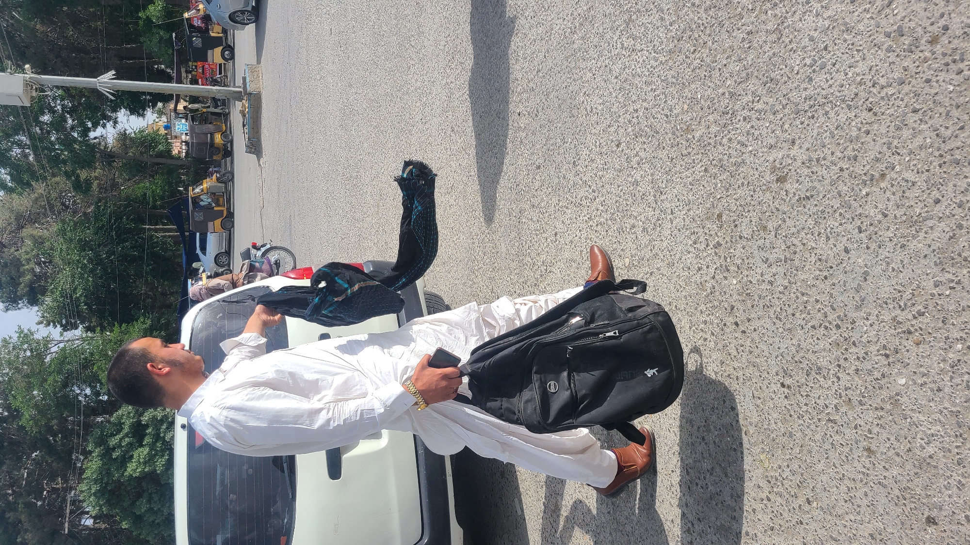 The image depicts a man standing next to a white car on a paved road in Quetta City Tehsil, Pakistan. The man is wearing a white shalwar kameez and brown shoes. He has a black backpack over his shoulder and is holding a black object in his hand. He is also holding a dark blue patterned cloth draped on the rear of the car. The background shows a street scene with trees, other vehicles, and buildings.