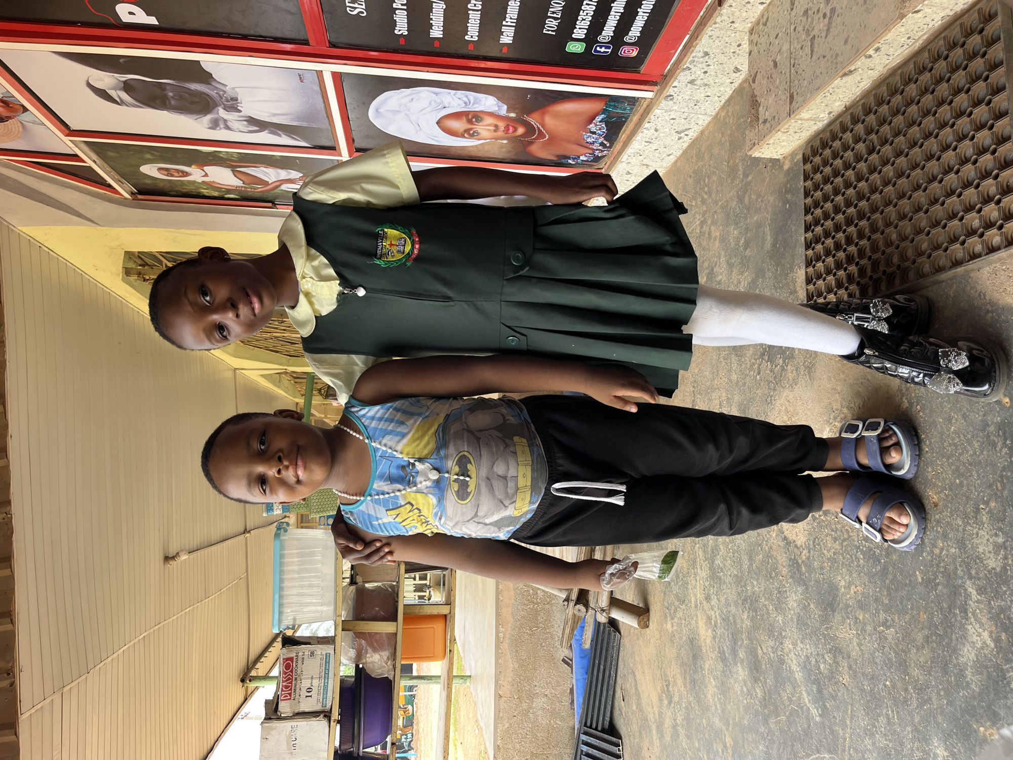 Two young children, a girl and a boy, are standing next to each other outdoors. The girl, on the left, is wearing a dark green school uniform with a light yellow collar and a school crest. She has dark hair and is looking directly at the camera with a neutral expression. The boy, on the right, is wearing a blue and yellow sleeveless t-shirt with a Batman design and black pants. He has dark hair and is also looking at the camera with a slight smile. The background shows what appears to be a shop or stall with framed pictures and shelves filled with various items, including what looks like cookware and plastic containers. Some text is visible on the framed pictures, advertising services like "Studio Portfolios," "Wedding/Event Photography," "Content Creation," and "Wall Frames." There is also a phone number and social media handles visible. The lighting suggests it is daytime. The children are standing on a textured outdoor floor, with a black mat with a circular pattern visible to the right of the girl. The overall scene appears to be a casual, everyday setting.