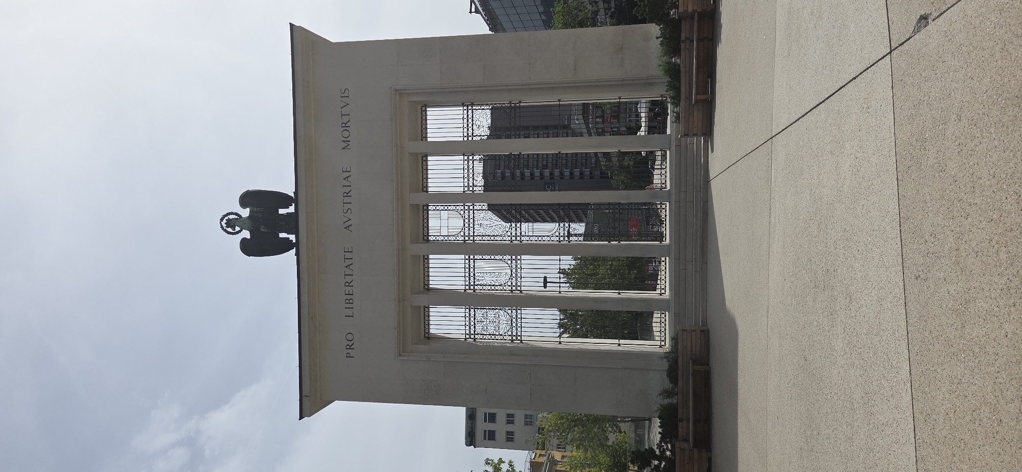 The image depicts the Innsbrucker Heldenorgel, a war memorial located in Innsbruck, Austria. The monument is a large, pale stone structure with an eagle statue at the top. Inscribed on the facade is the Latin phrase "PRO LIBERTATE AVSTRIAE MORTVIS," which translates to "For Austria's freedom, the dead." The monument features a grand entrance with ornate metal gates. In front of the monument, a paved plaza is visible, with wooden benches and planters. Behind the monument, modern buildings and trees can be seen, indicating an urban setting. The sky is partly cloudy. There are no people or animals in the frame.