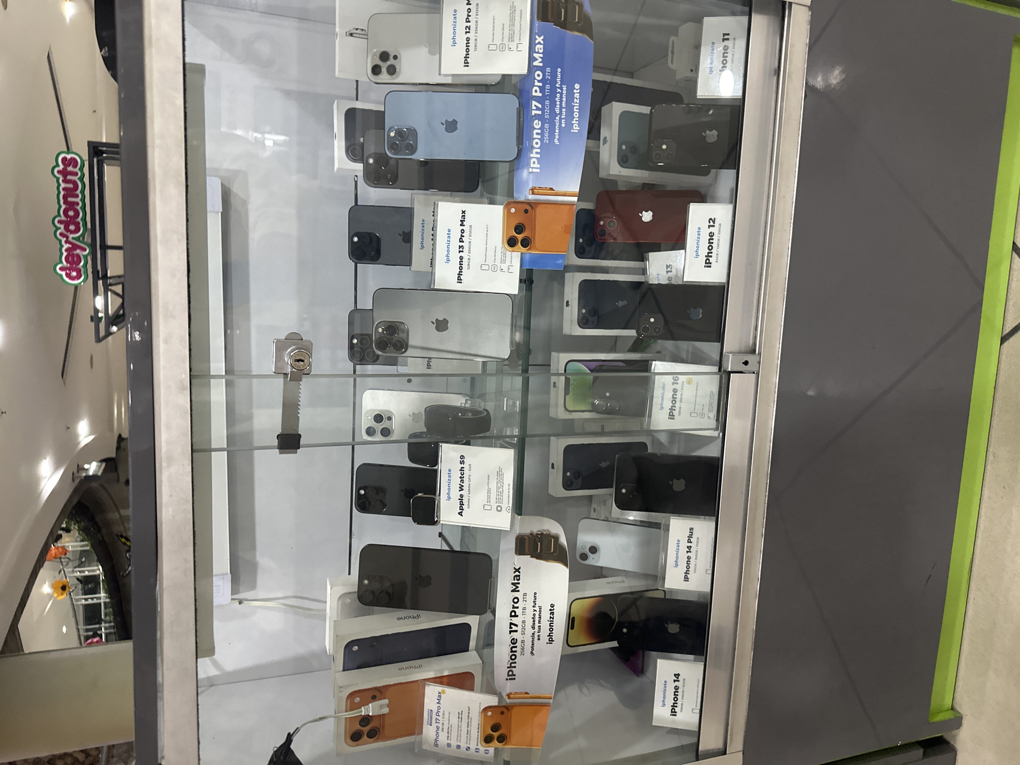 The image shows a display case of various iPhones for sale, along with an Apple Watch. The phones range from iPhone 11 to iPhone 17 Pro Max. There is signage within the case indicating that the phones are being sold by "iphonizate". In the reflection of the glass, a "dey'donuts" sign is visible. There is no explicit evidence in the image to determine that the location is in the city of Caracas, Venezuela.
