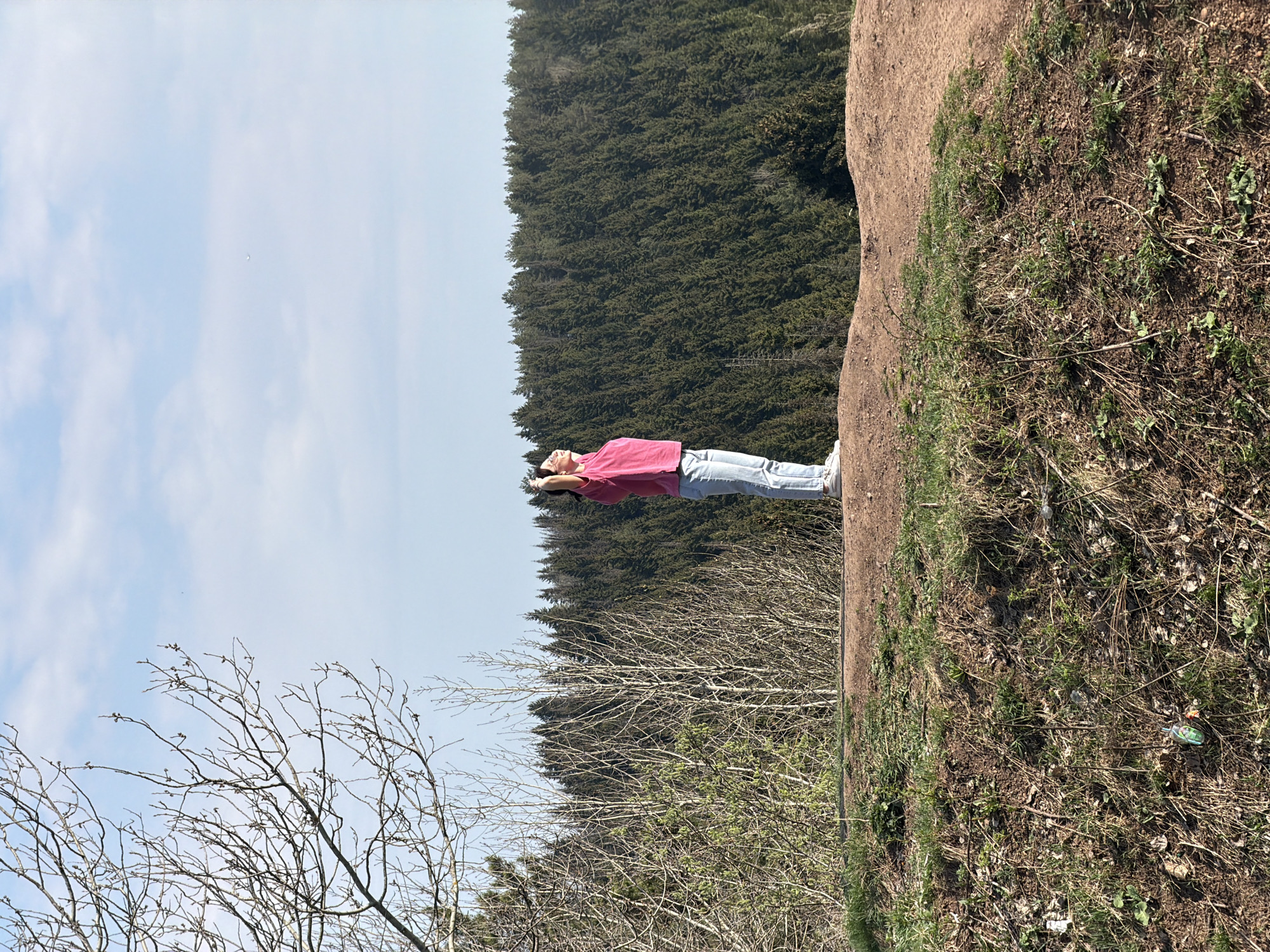 A medium, slightly upward angled shot shows a young woman standing on an earthen cliff edge, arms raised and head tilted back. She is wearing a pink long-sleeved shirt, light blue jeans, and white sneakers. Behind her is a dense forest of evergreen trees. The sky is visible above the trees, with some wispy clouds. In the lower left of the frame are bare trees with emerging buds, indicating spring. The ground is brown and somewhat barren, with some green shoots. The location is a cliff edge, possibly near Perm, Russia. The woman appears to be in a state of enjoyment or relaxation, with her arms raised toward the sky.