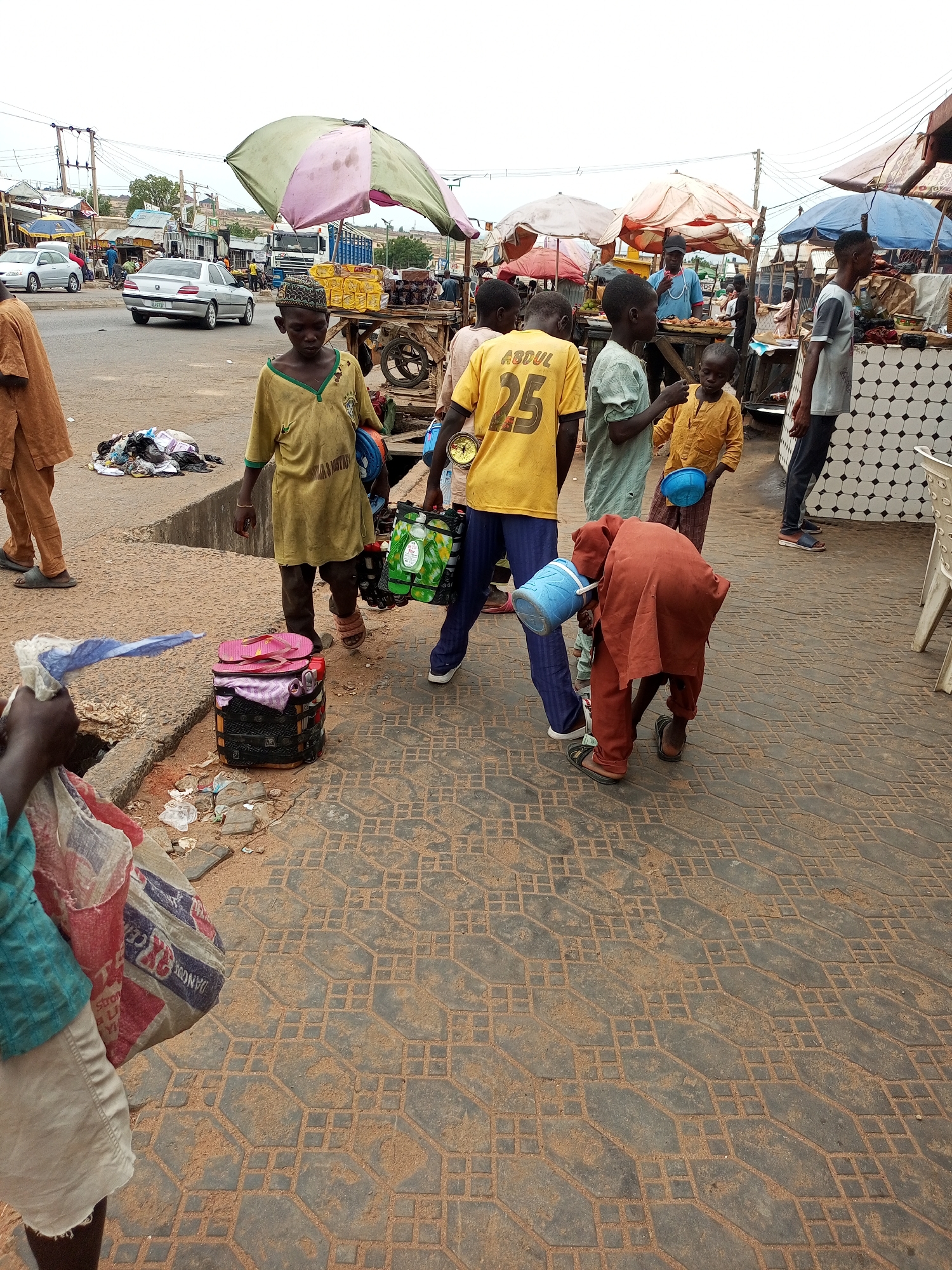 A wide shot captures a bustling street scene in Gombe, Nigeria, likely a market area based on the presence of vendors and shoppers. A young boy in a yellow t-shirt with "ABDUL 25" on the back stands near a market stall. To his left, another boy in a green shirt holds a blue bucket, and to his right, a boy in a yellow kaftan also carries a blue bucket. In the foreground, a person in an orange robe bends down, holding a blue bucket. Behind them, a few adults and children are seen in the background, some interacting with market stalls. A silver car is parked on the street, and a pile of clothing lies on the ground near the curb. The ground is paved with interlocking stones, and there are various market umbrellas and stalls scattered throughout the scene. The sky is overcast, suggesting a cloudy day.