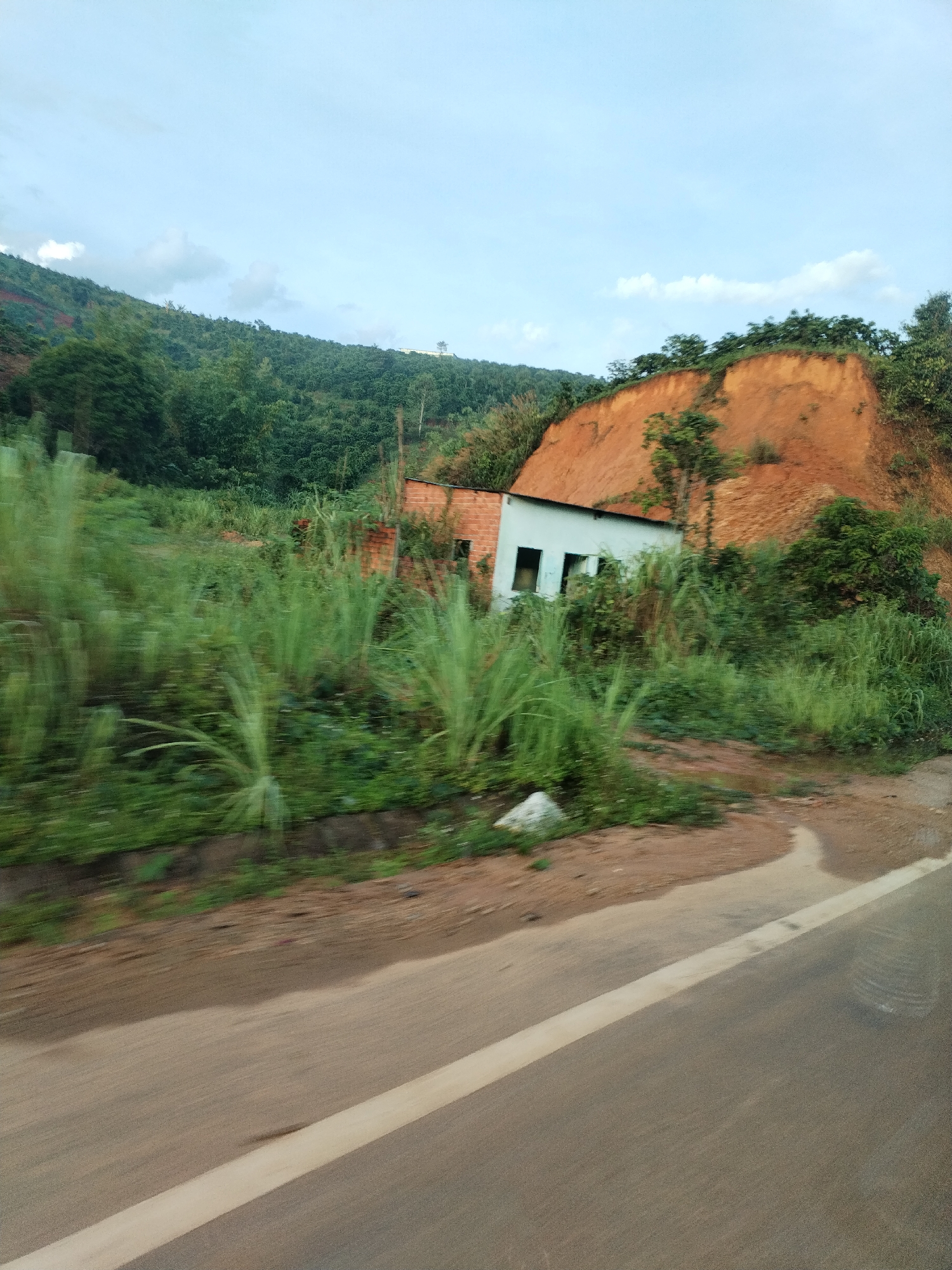 A blurry, eye-level photograph depicts a roadside scene in Bo Y, Vietnam. The main subject is a partially obscured building nestled against a red clay hillside. The building has sections of exposed brick and white siding with what appear to be window openings. Overgrown vegetation, mainly tall grasses, dominates the foreground and obscures much of the building. The hillside, composed of reddish-brown earth, is partially covered with trees and foliage along its crest. Behind the hillside, a forested mountain range rises into a sky with scattered clouds. The bottom portion of the image reveals a section of asphalt road with a solid white line marking the edge, suggesting the photograph was taken from a moving vehicle. The blur indicates movement and a rapid shutter speed.