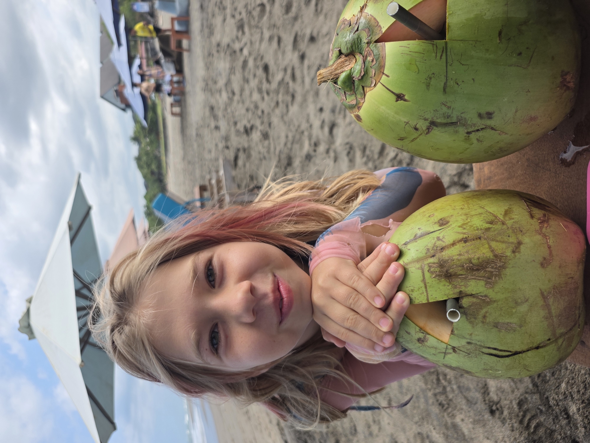 A young girl with blonde hair and pink highlights, wearing a pink and blue top, is pictured leaning on a table on a beach in North Kuta, Indonesia. Two green coconuts with straws inserted sit on the table. In the background, there is sand, vegetation, and beach umbrellas with people seated underneath. The sky is visible in the upper left corner.