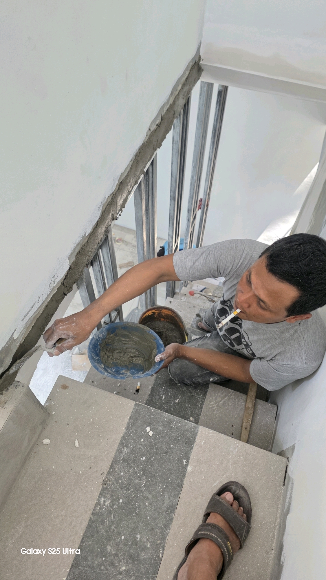 The image depicts a man working on what appears to be a construction site, likely indoors given the presence of walls and what seem to be building materials. The man, wearing a gray t-shirt and dark pants, is seated on a staircase and appears to be applying plaster or cement to a wall with a trowel. He is also holding a blue bucket filled with the same material. A cigarette is visible in his mouth, suggesting he is taking a break or working while smoking. Another orange bucket is partially visible behind him. The staircase is made of stone tiles with a checkered pattern, and there are metal framing elements visible in the background, suggesting ongoing construction. The overall environment suggests a residential or commercial building under renovation or construction. There is no specific indication of the location within Juwana, Indonesia, as no identifying landmarks or signs are present.