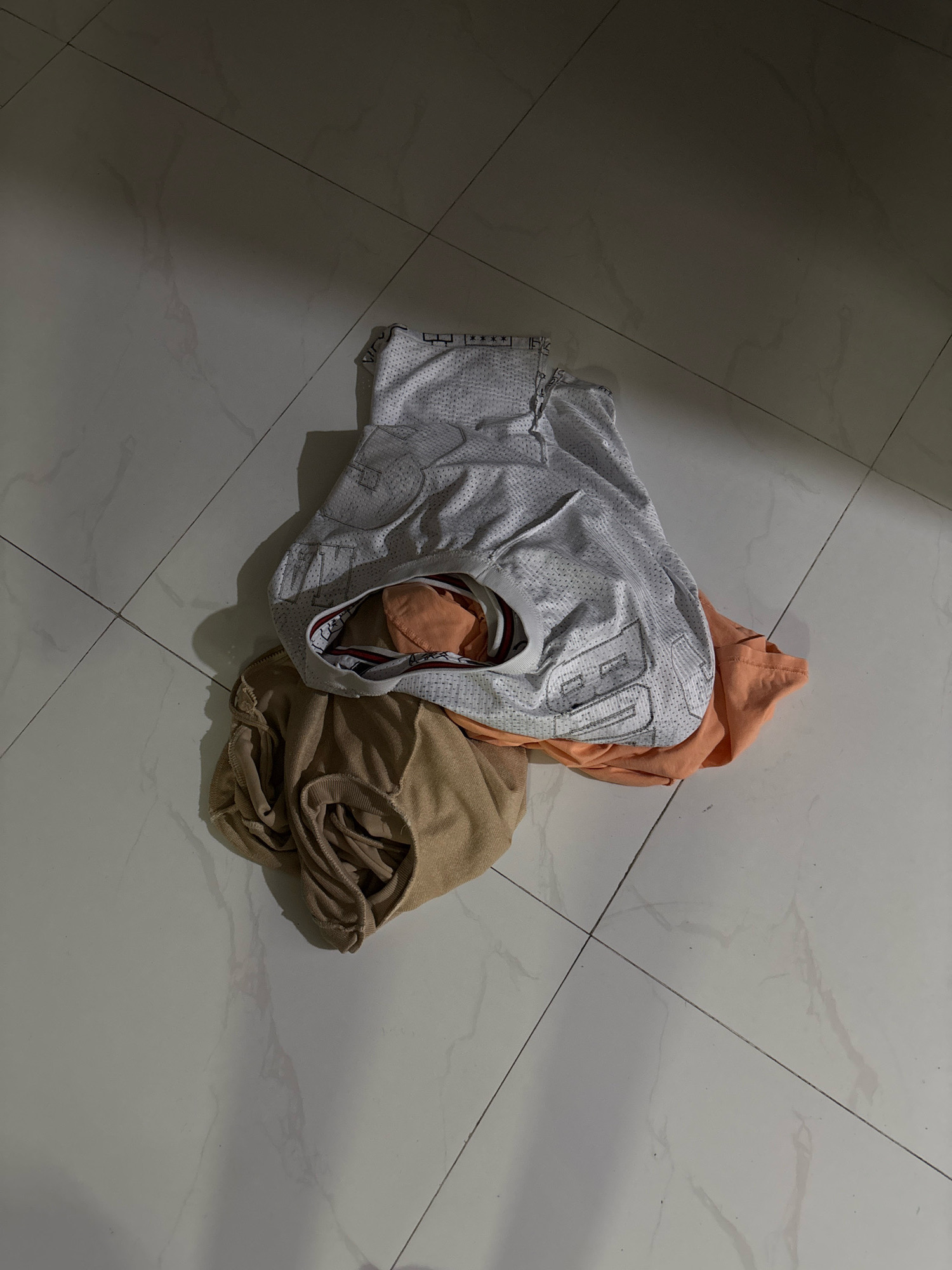 A pile of dirty laundry sits on a light-colored tiled floor. The laundry consists of a light gray mesh shirt with "85" printed on it, an orange t-shirt, and a tan garment. The lighting suggests an indoor setting, possibly a bathroom or bedroom. The tiles are large and have a marble-like pattern. There is a shadow cast from the left side of the image, indicating a light source. The scene is devoid of people and any signs of struggle or disarray beyond the scattered clothes. There are no discernible features that would place the scene in Nha Trang, Vietnam.