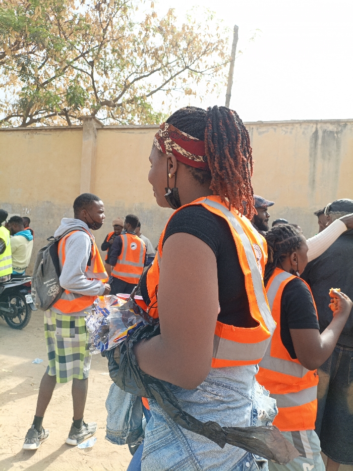 The image depicts an outdoor gathering in Sabon Gari East, Nigeria, featuring multiple individuals, many wearing orange high-visibility vests with reflective stripes, suggesting a community activity. In the foreground, a female subject is positioned with her back partially to the viewer, turned slightly to her left. She wears a black short-sleeved top, light-wash denim bottoms, an orange high-visibility vest featuring a circular logo on the back, and a patterned red and white headband. Her hair is styled in dark dreadlocks or braids, pulled back into a ponytail with reddish-brown tips. A black face mask hangs from her left ear. She holds a black plastic bag containing clear plastic items, possibly bottles or wrappers, under her left arm.
To her left, in the mid-ground, a male subject faces right, looking towards the foreground female. He wears a grey hooded sweatshirt, green and white plaid shorts, black sneakers, a black backpack, and an orange high-visibility vest. Visible on the ground near him are discarded plastic items. Further left, the rear wheel and frame of a motorcycle are partially visible. In the mid-ground to the right of the foreground female, another female subject in a black top and orange high-visibility vest is seen eating a yellow or orange food item with her right hand.
Several other individuals are visible in the background, many also in orange high-visibility vests, indicating a coordinated group. The environment includes a light-colored sandy ground, a tall, plain beige wall, and trees with sparse, light-green to brownish foliage under a bright sky.