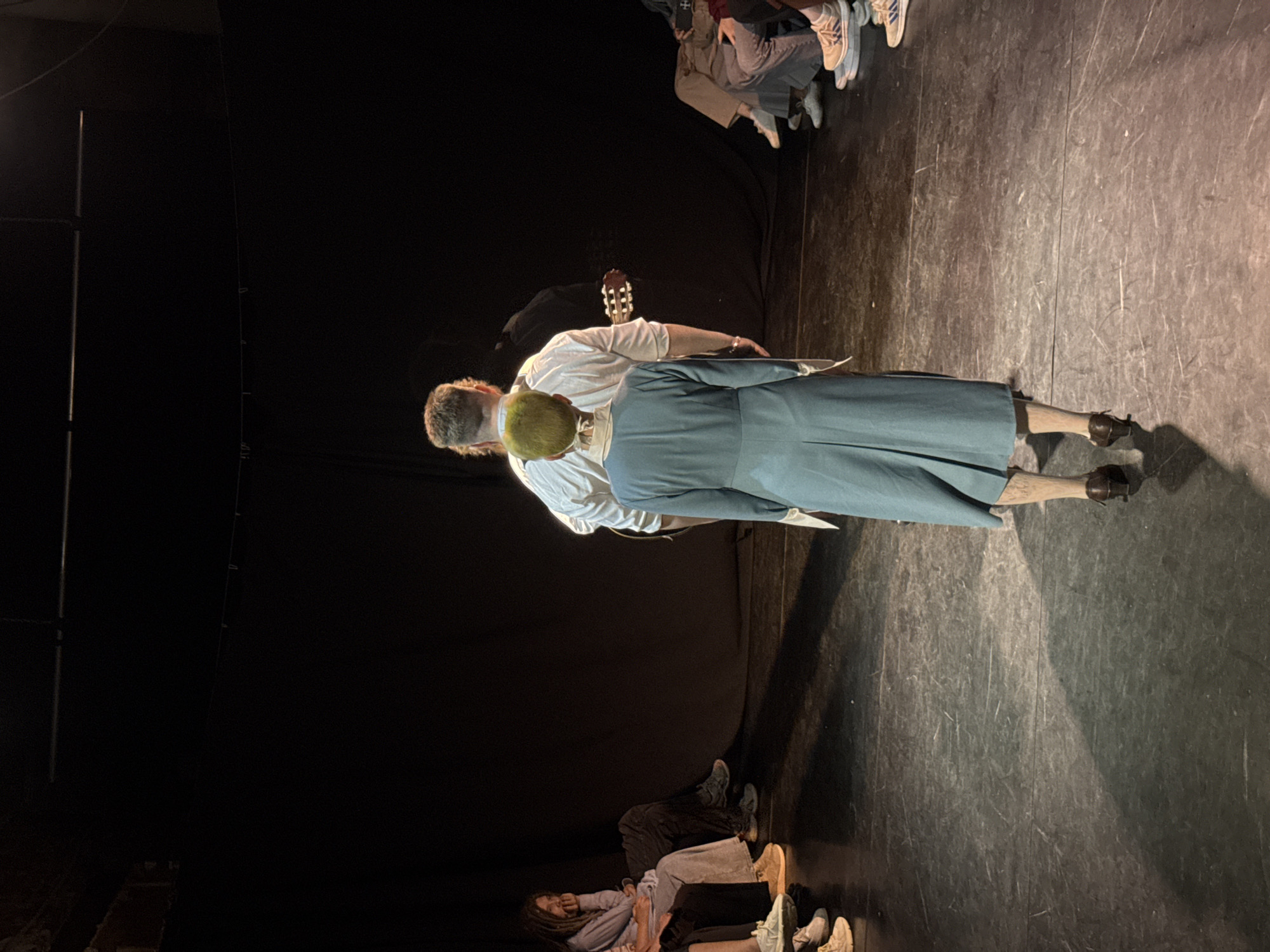 The image captures a scene on what appears to be a stage, likely in a theatre. Two individuals are centrally positioned: one wearing a white shirt and carrying a guitar, and the other in a long, teal-colored dress or coat. The latter has noticeable yellow hair. They seem to be engaged in a performance or rehearsal. Several people are sitting on the floor and watching the event. The environment includes a black backdrop. The stage floor is dark and textured. Given the mention of the city of Izhevsk, Russia, the location is probably a theatre in Izhevsk.