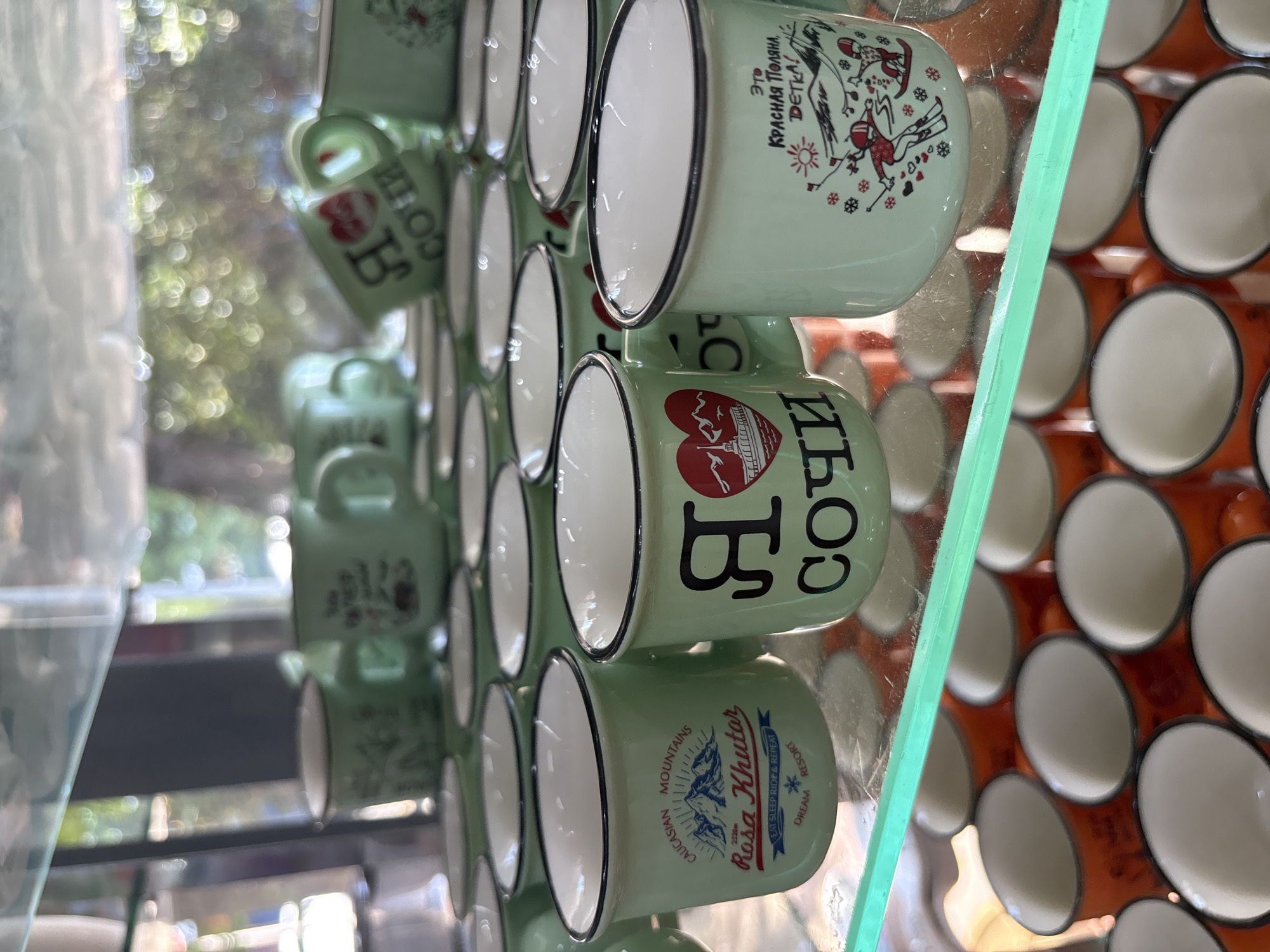 The image displays a collection of small, light green enamel mugs arranged on shelves within a display case, likely in a souvenir shop in Sochi, Russia. Several mugs feature designs related to the city and its surroundings. One mug prominently shows "Caucasian Mountains" and "Rosa Khutar" with an image of mountains and skiers. Another mug has a stylized heart emblem with a building and the text "Я" and "СОЧИ" (which translates to "I love Sochi"). A third mug depicts skiers on a snowy slope with the text "Это Красная Поляна, Детка!" (This is Krasnaya Polyana, baby!). The mugs are neatly organized, with some stacked behind others. The background appears to be an indoor setting with a blurred view of greenery and sunlight, suggesting the shop is near an outdoor area. Below the green mugs, a layer of orange mugs is visible, also arranged in rows. The scene captures a typical retail display of tourist merchandise.