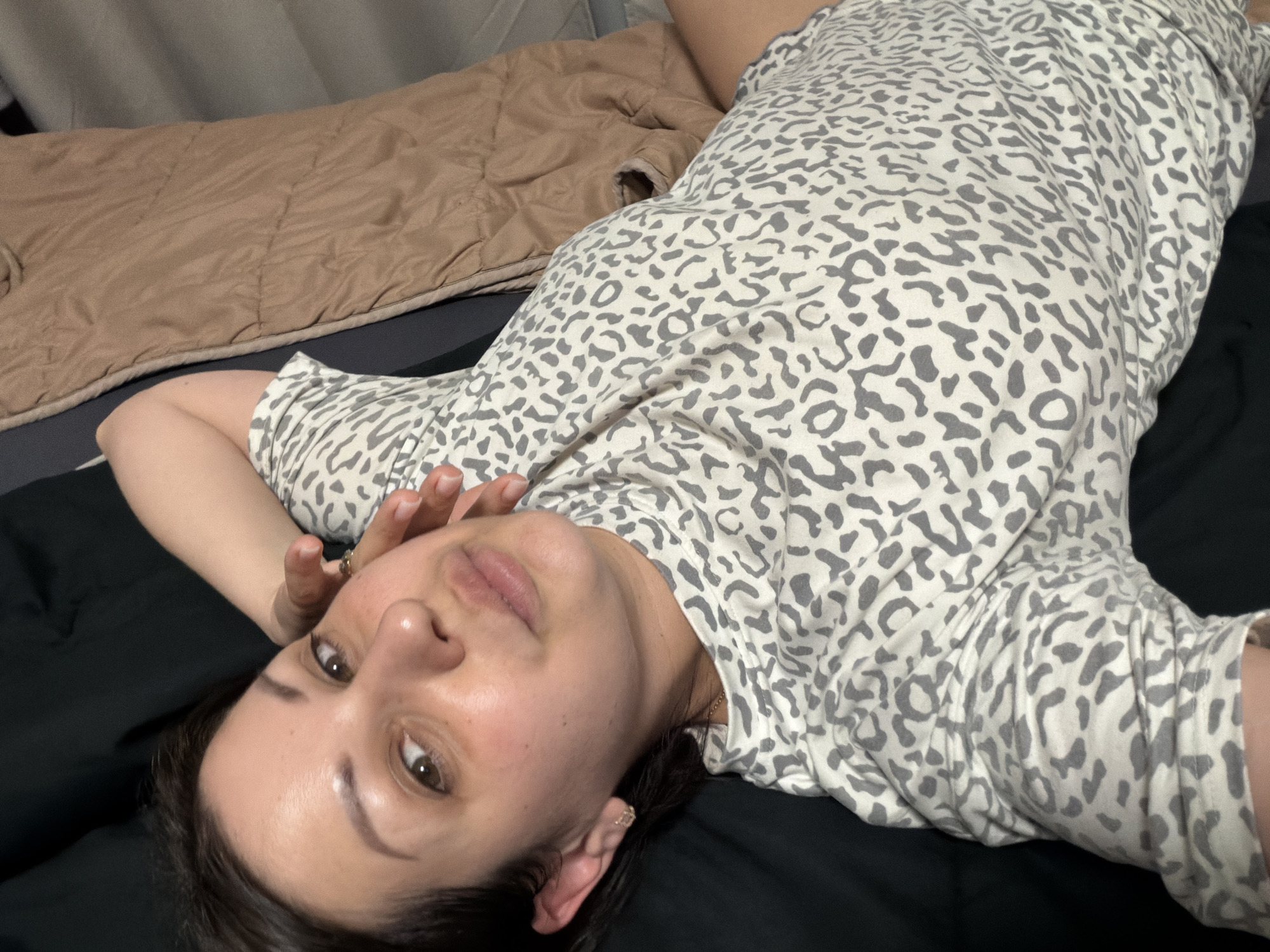 A woman with dark hair is lying on her back on a bed, looking up at the camera. She is wearing a grey and white leopard print top. Her left arm is bent, with her hand resting on her face. She has light skin and brown eyes. The bed is covered with a brown quilted blanket and a black sheet. The scene appears to be indoors, possibly in a bedroom. The lighting suggests it could be daytime or a well-lit room. There is no visible text in the image. The overall mood seems relaxed and casual.