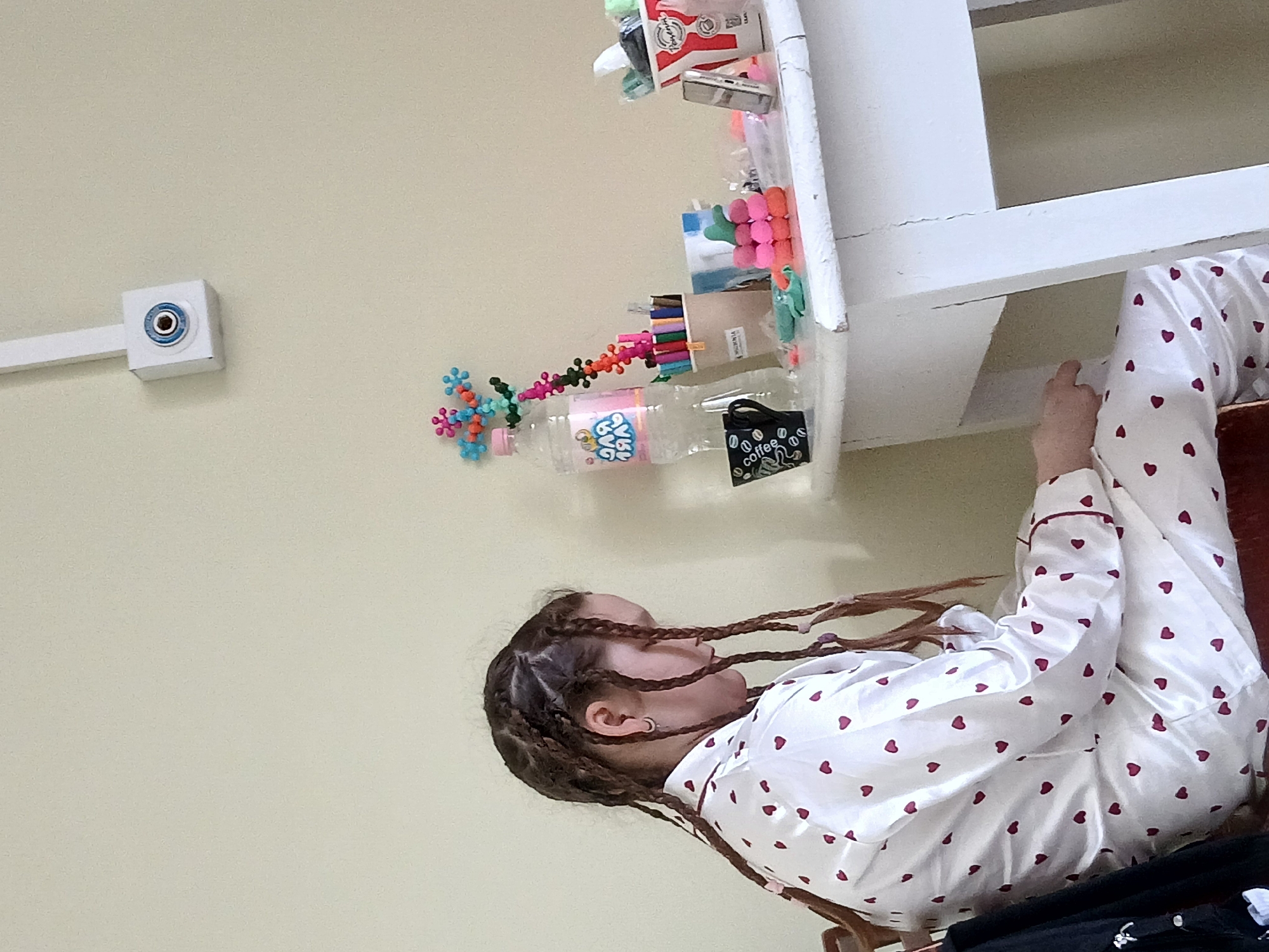 A bedroom interior features a young woman in white pajamas patterned with red hearts, sitting with her legs resting on a white shelf cluttered with various items, including a bottle labeled "BUG BUG." She has numerous thin braids extending from her head. The background is a plain beige wall with a small, square white device attached to it.