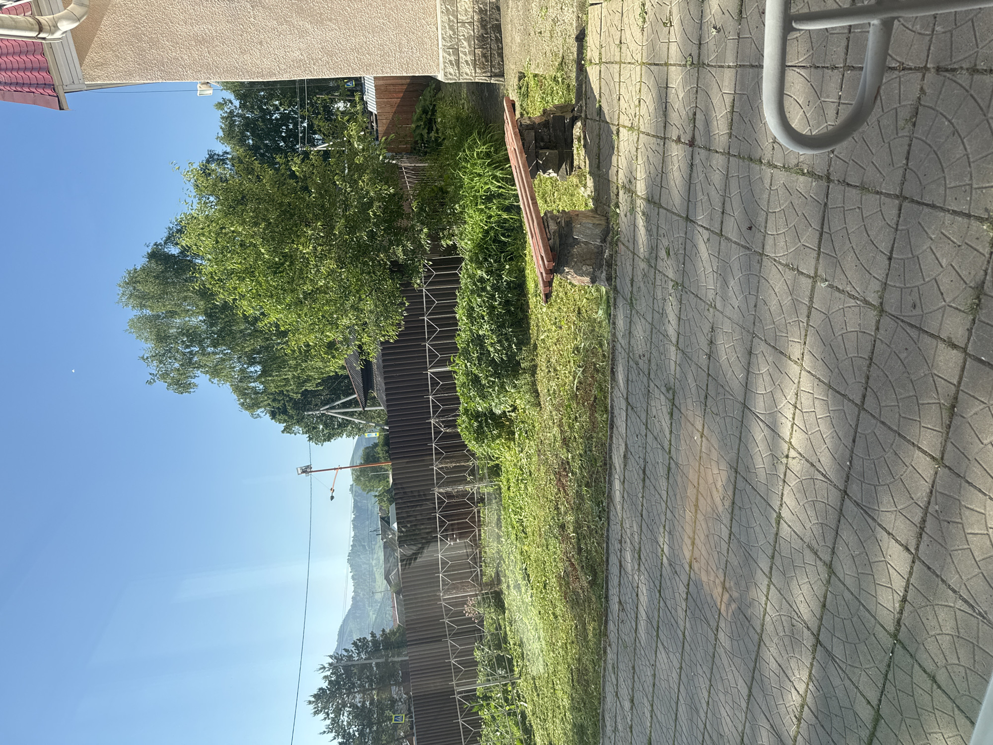 The image depicts a sunny outdoor scene with a tiled pathway in the foreground. To the right, a light-colored building with a red roof is partially visible. In the middle ground, a simple bench made of stone supports and wooden planks sits on the pathway. Behind the bench, a lawn with recently cut grass stretches towards a dark wooden fence. Beyond the fence, lush green trees and vegetation are visible, with a distant view of rolling hills and mountains under a clear blue sky. Power lines and a street lamp can be seen in the background. The overall impression is a quiet, residential area on a bright day.