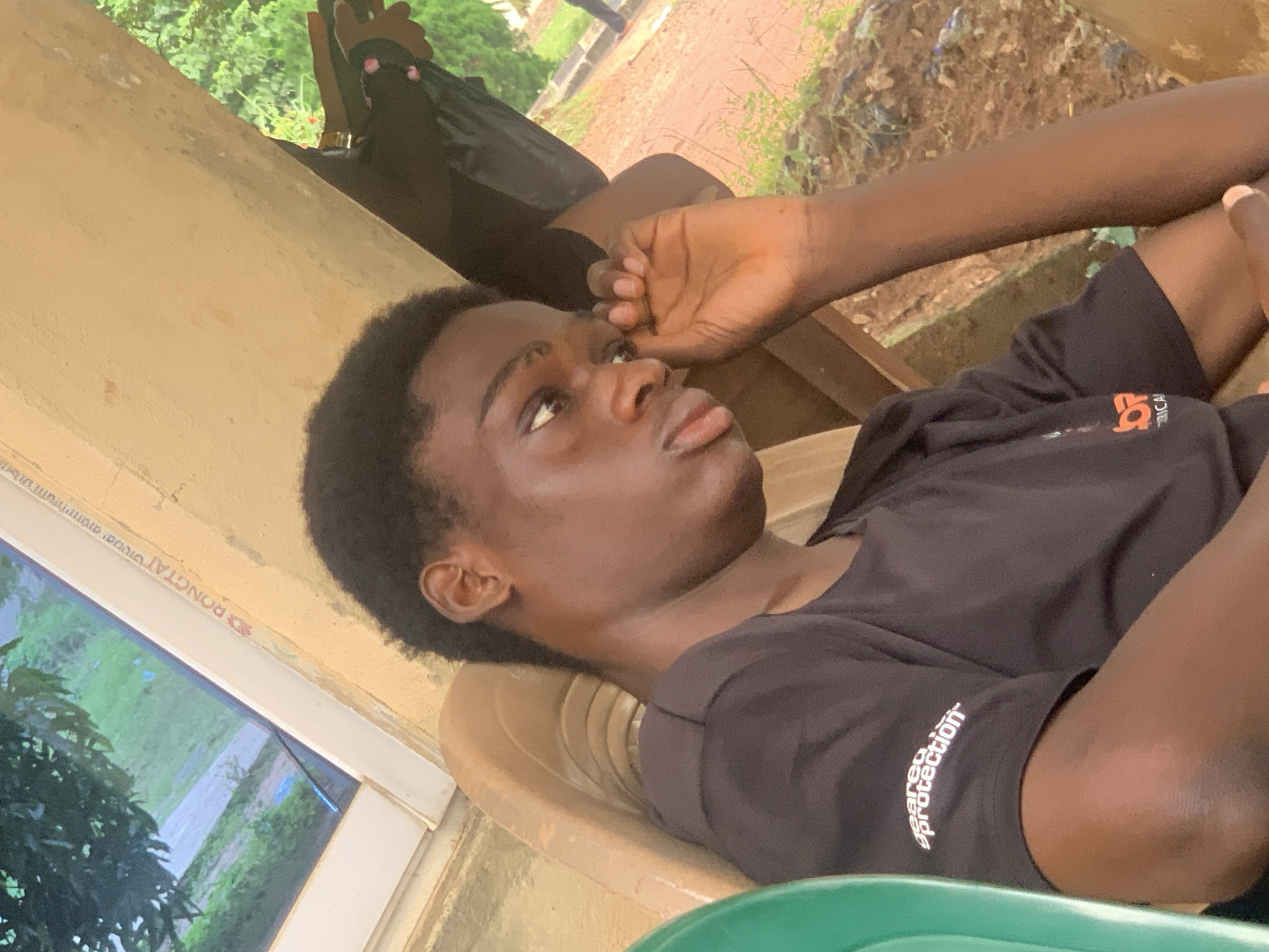 The image shows a young Black male sitting in an outdoor setting in Ukukwa, Nigeria. He is wearing a black t-shirt with white text that reads "geared protection". His right hand is raised to his face, with his fingers touching his eye. The background consists of a yellow-painted wall and a glimpse of greenery, suggesting a warm climate. The man is seated in a light-colored plastic chair. The overall scene appears casual and relaxed, with the man perhaps tired or thoughtful.