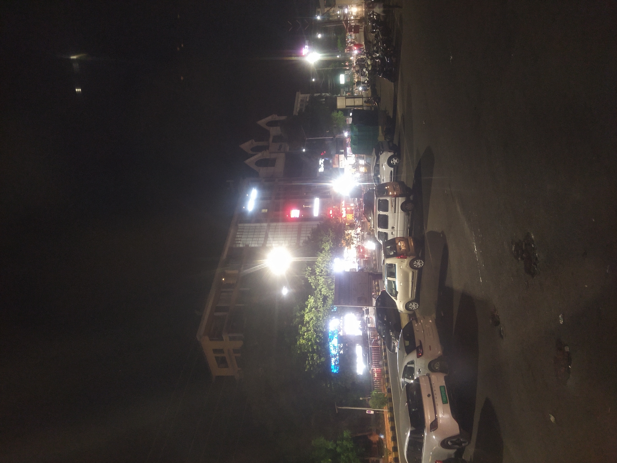 Captured at night in Margao, India, the image presents a congested street scene illuminated by streetlights and building signage. A row of parked cars lines the roadside, ranging from white compact vehicles to darker sedans. Pedestrian activity is suggested by the presence of motorbikes parked along the edge of the road. The cityscape is visible in the background, defined by tall buildings and bright lights, suggesting commercial activity. The overall atmosphere is urban and active, indicative of a bustling city environment.