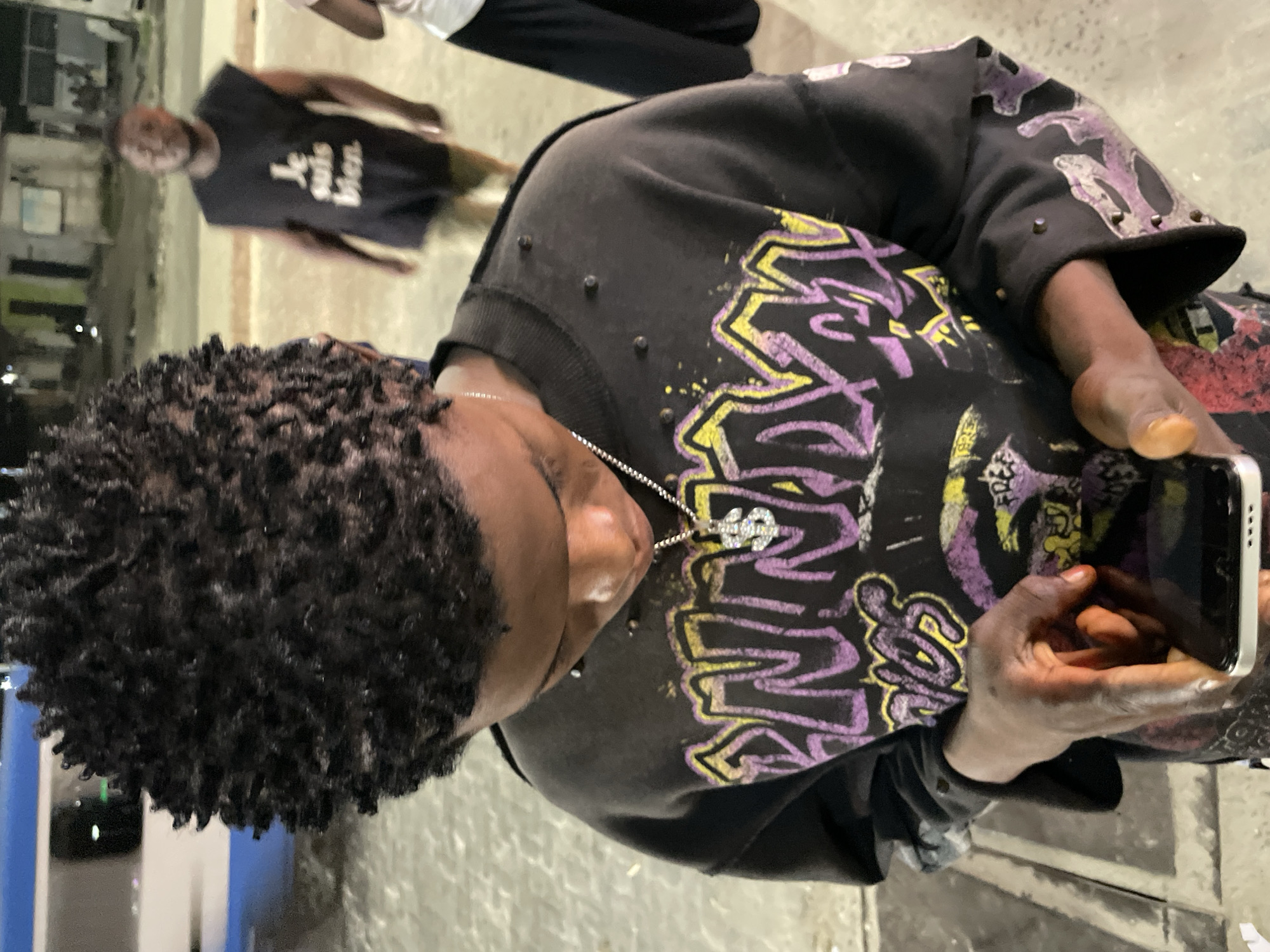 A medium shot captures a young Black male from the chest up, looking down at a smartphone in his hands. He has short, dark, coiled hair and is wearing a black crewneck sweatshirt adorned with a vibrant, graffiti-style graphic in purple and yellow. A silver chain with a dollar sign pendant rests on his chest. The background is dimly lit, suggesting an outdoor urban setting at night. In the mid-ground, to the right of the primary subject, another Black male stands facing away from the camera, wearing a black sleeveless t-shirt with white text that reads "Je suis bien." To the far right, partially visible, is a third individual wearing a white top and black pants. The environment appears to be paved with concrete, and a blurred blue and white object, possibly a vehicle or bus, is visible in the far left background. The overall impression is a candid street scene in Ikorodu, Nigeria.