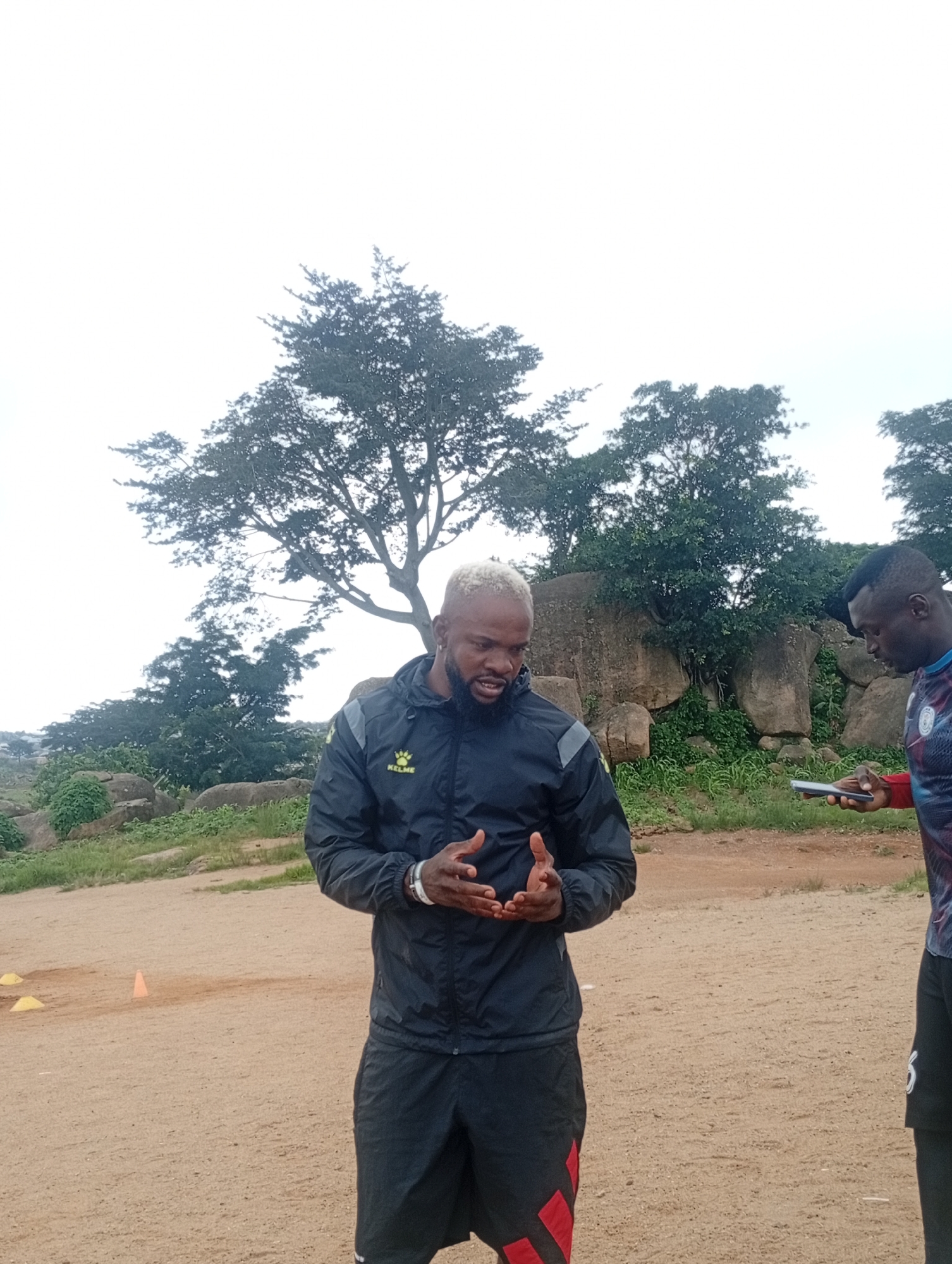 Two Black men are standing outdoors on a sandy surface, likely a sports field. The man on the left has bleached blonde hair and a beard and is wearing a black athletic jacket with grey accents and black athletic shorts with red stripes. He is facing the viewer and gesturing with his hands, as if speaking. The man on the right, with dark hair, is wearing a multi-colored athletic shirt and black shorts. He is looking down at a smartphone he is holding in his left hand. In the background, there are large rocks and trees, suggesting a somewhat rugged natural environment. The sky is overcast. The scene appears to be in Jos, Nigeria, given the context of the image's origin.