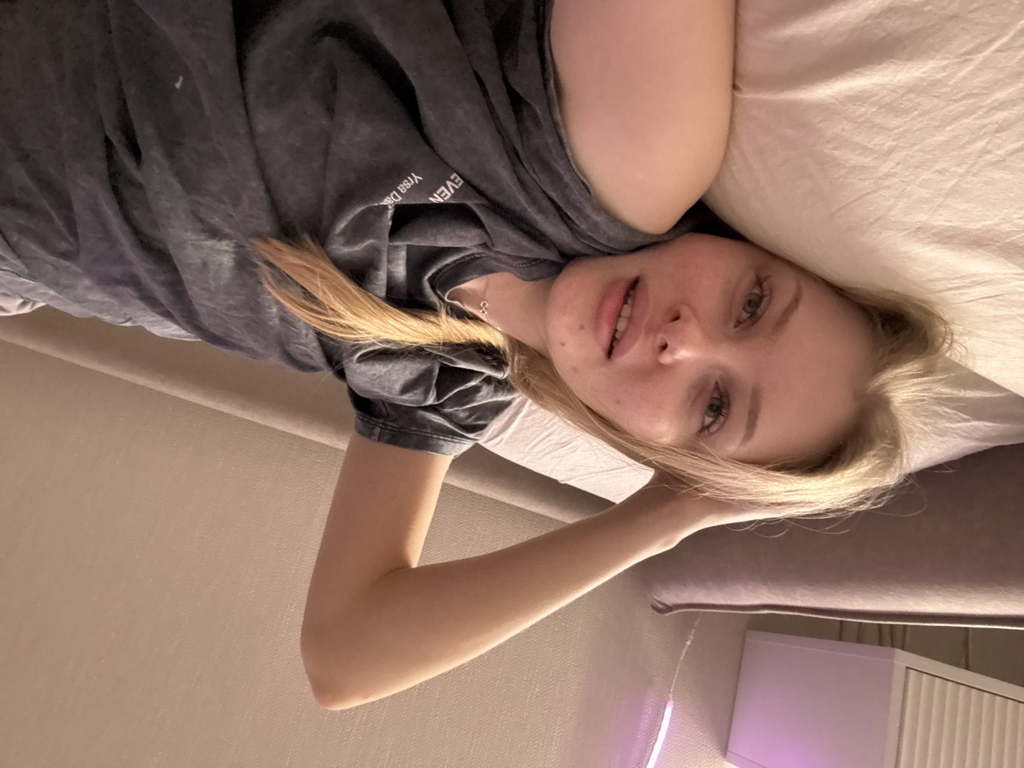A young, fair-skinned woman with blonde hair is lying on a bed. She is wearing a grey t-shirt. Her head rests on a white pillow. Her left arm is bent and raised, with her hand near her head. A section of a wall with a beige tone is visible to the left of the bed. A purple and white object, potentially a lamp or light fixture, is partially visible in the lower right corner of the frame. The photo appears to be taken indoors, and there is no information available to determine the location as Perm, Russia.