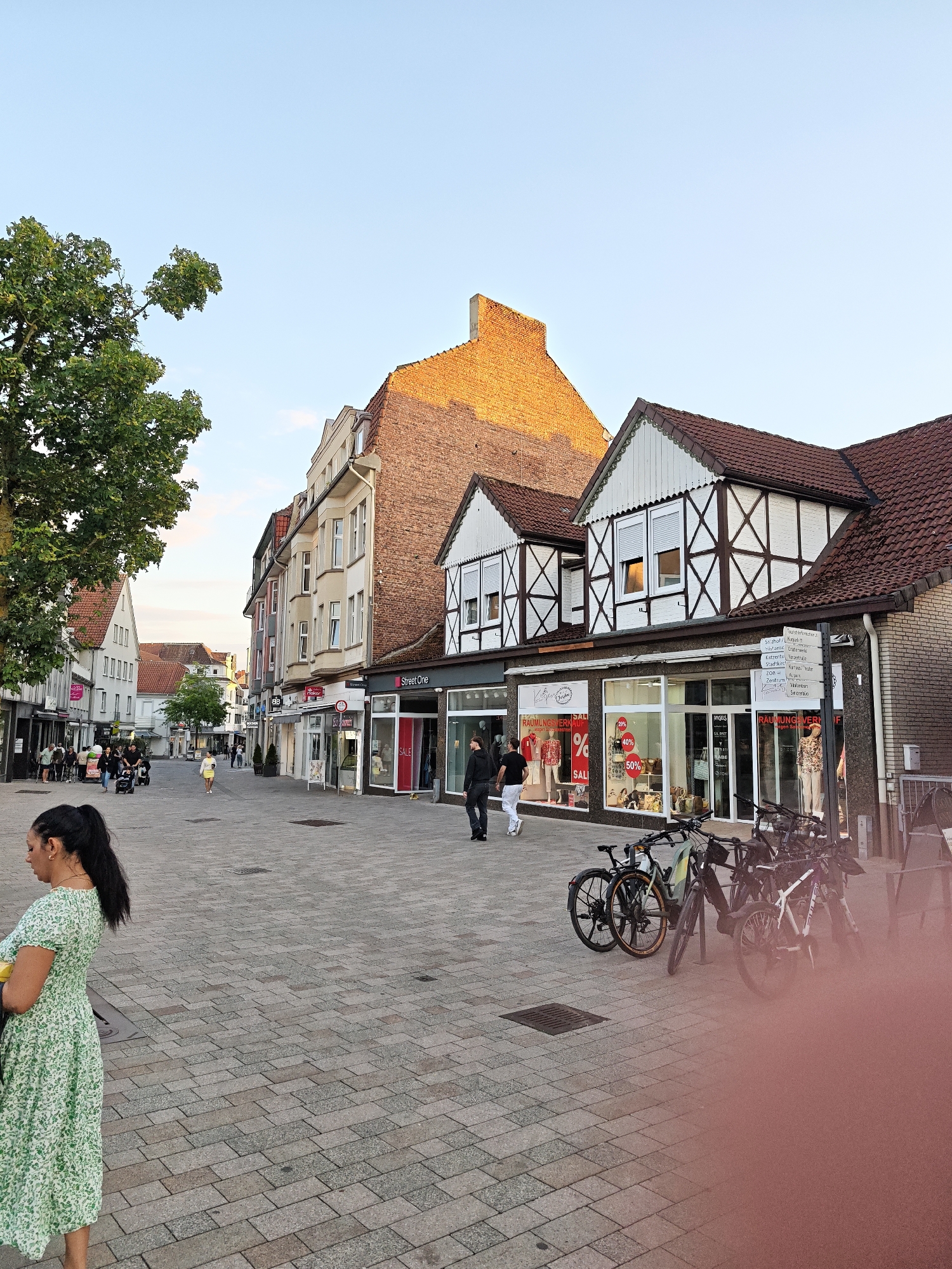 The image depicts a street scene in Bad Salzuflen, Germany, specifically the pedestrian zone. On the right side of the street, several shops are visible, including "Street One" and "Raumung Verkauf," the latter featuring a mannequin display and sale signs indicating 20%, 40%, and 50% discounts. A signpost with multiple directional arrows is also present. Several bicycles are parked along the sidewalk in front of the shops.
On the left side of the street, more shops and buildings line the pedestrian area. A large tree with green foliage stands prominently in the foreground on the left. Several people are walking along the street, including a woman in a green floral dress in the foreground, a couple of people pushing a stroller, and other individuals browsing or walking. The buildings exhibit a mix of architectural styles, with some having traditional timber framing and others appearing more modern. The street itself is paved with cobblestones. The sky is clear and blue, suggesting daytime, possibly late afternoon or early evening due to the lighting.