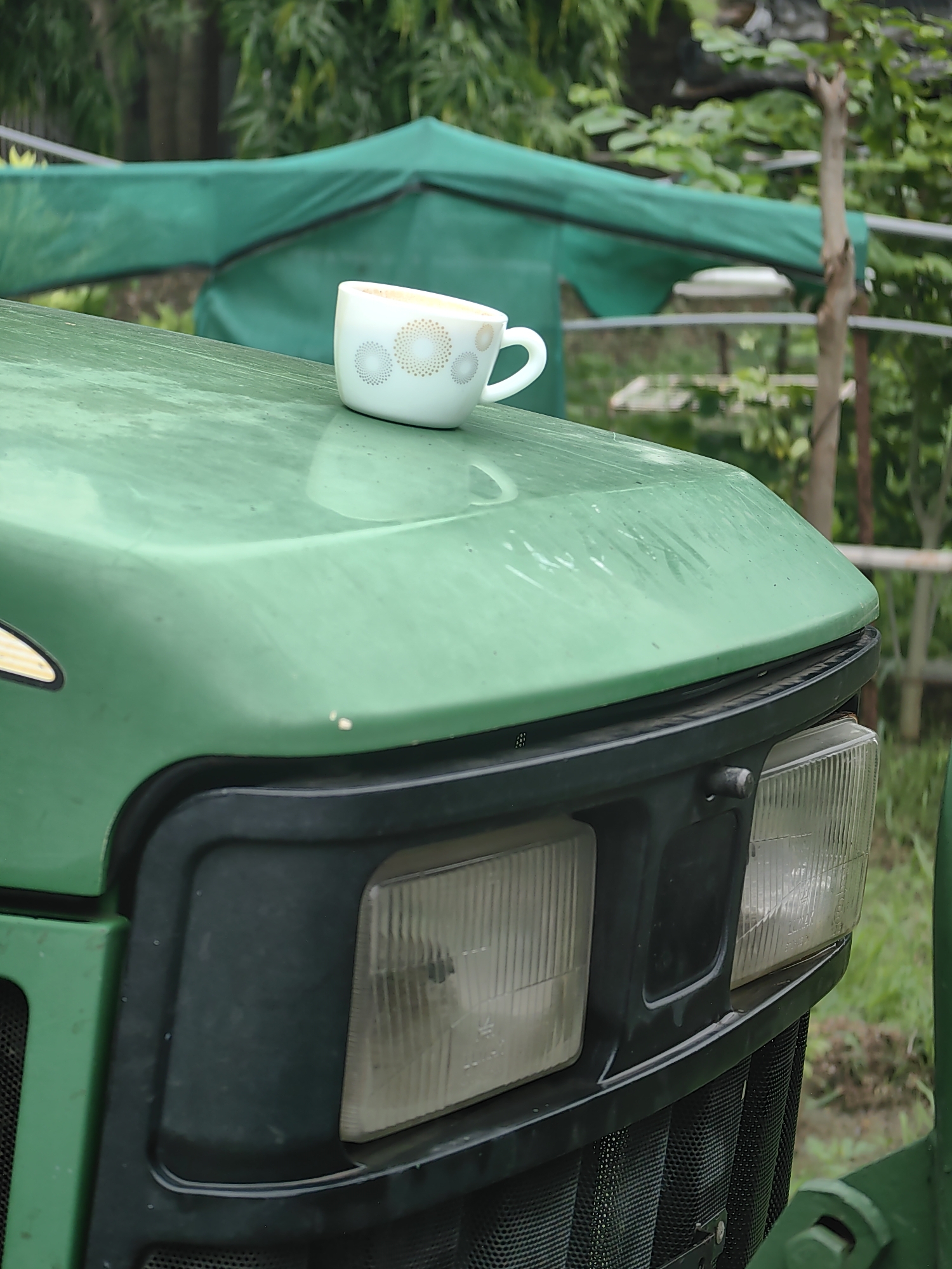 The image shows a white coffee cup with a subtle decorative pattern of concentric circles in gold and grey, placed on the green hood of a tractor. The tractor's headlights are visible in the foreground. In the background, a green tarp or shade structure is erected, with lush green foliage and trees beyond. The scene appears to be outdoors, likely in a rural or agricultural setting in Mhaiswadi, India, suggested by the presence of the tractor and the dense vegetation. There are no people visible in the image. The lighting suggests it might be an overcast day.