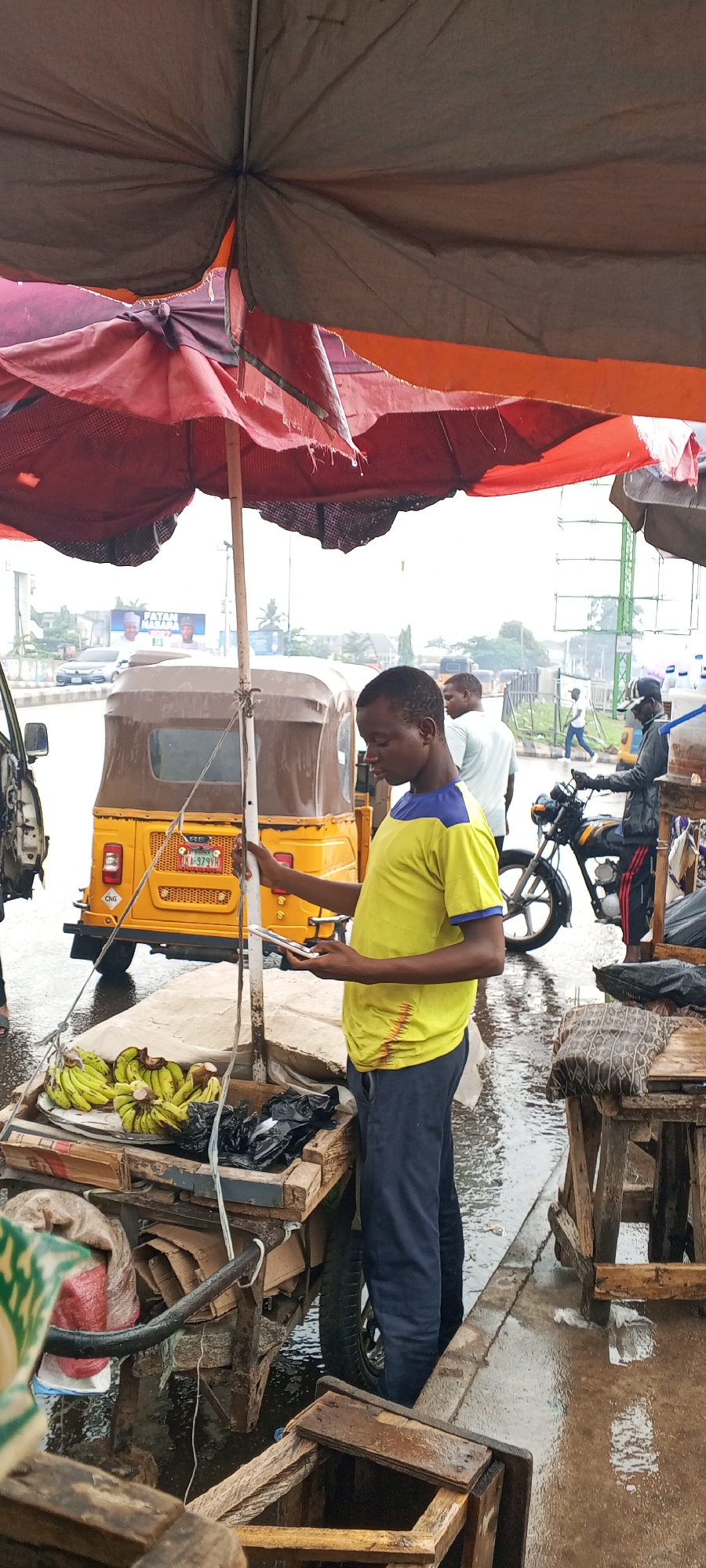 The image captures a street scene in what appears to be Maiburiji, Nigeria, based on the visible features. A young man, dressed in a yellow shirt with blue sleeves and dark pants, stands beside a wooden cart laden with bananas and some goods in black plastic bags. He is looking down at his phone. The cart is under a partially torn, multi-layered sunshade. In the background, there is a yellow "keke," a type of auto-rickshaw common in Nigeria. Further back, people can be seen walking, and a motorcycle is parked nearby. The road and pavement appear wet, suggesting it has recently rained. Wooden stools or crates are visible at the edge of the frame. Overall, the image depicts a typical scene of informal commerce and transportation in a Nigerian urban area.