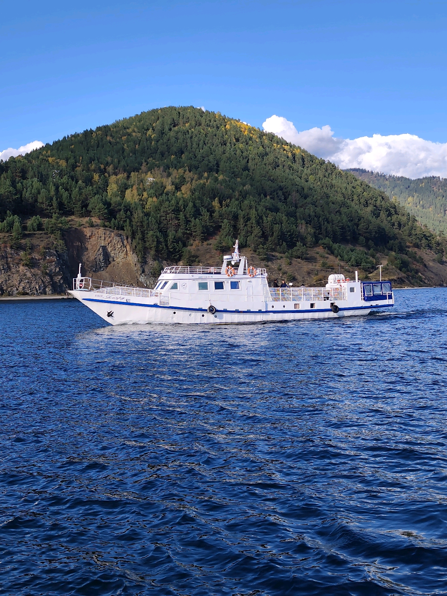 A white passenger boat with blue trim navigates on a deep blue body of water. The boat has multiple windows and railings, and several people can be seen on its deck. In the background, a steep, forested hill rises from the water's edge. The hill is covered in dark green coniferous trees, with patches of yellow and orange foliage indicating autumn. The sky above is a clear blue with scattered white clouds. The scene appears to be set in Listvyanka, Russia, likely on Lake Baikal, given the characteristic landscape.