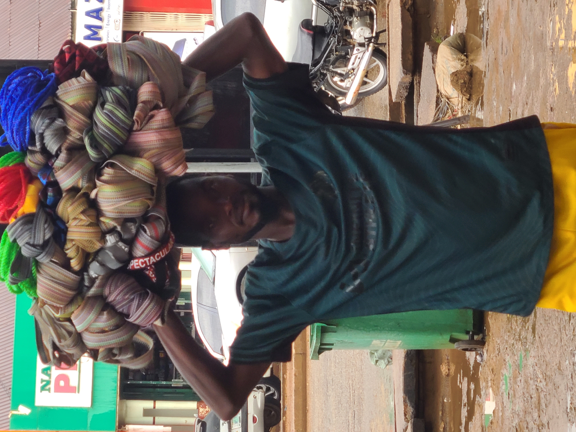 A Black man stands with his arms raised, balancing a large, colorful pile of rolled fabrics and ropes on his head. He wears a dark green t-shirt and bright yellow pants. The man is positioned outdoors, with a green trash bin to his left. In the background, a motorcycle is parked. The ground is wet and muddy, with puddles of water and debris scattered around. Behind the man, shop signs are visible, one reading "NA P" and another with "MA". The scene appears to be in a commercial area of Jos, Nigeria, indicated by the shops and the common sight of individuals carrying goods on their heads.